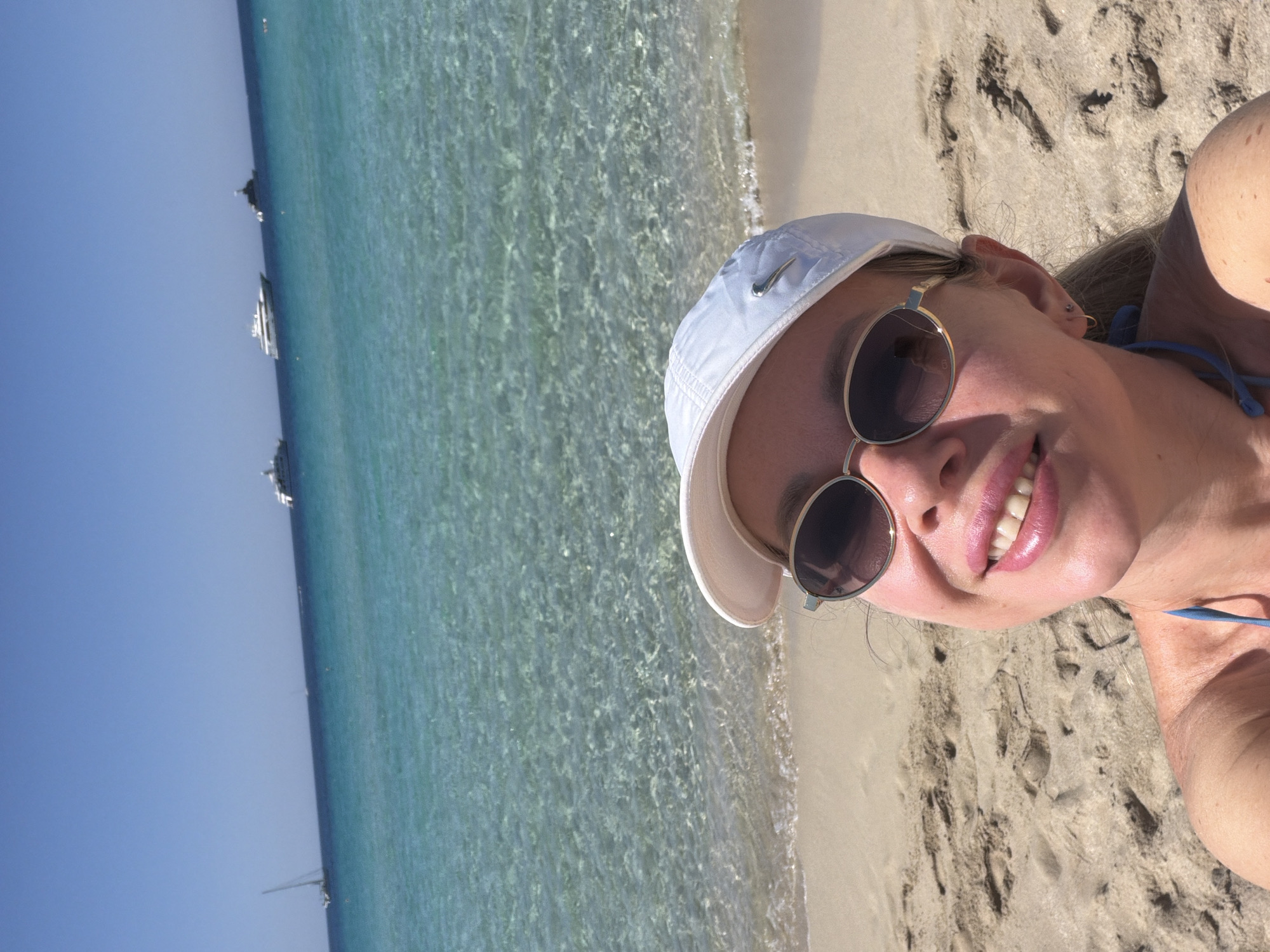 A woman wearing a white baseball cap and sunglasses smiles at the camera on a sandy beach. The ocean, with clear turquoise water, is in the background, with several yachts visible on the horizon. The sky is a clear, bright blue. The woman has fair skin and is wearing a blue bikini top. The overall scene depicts a sunny day at a beach location.