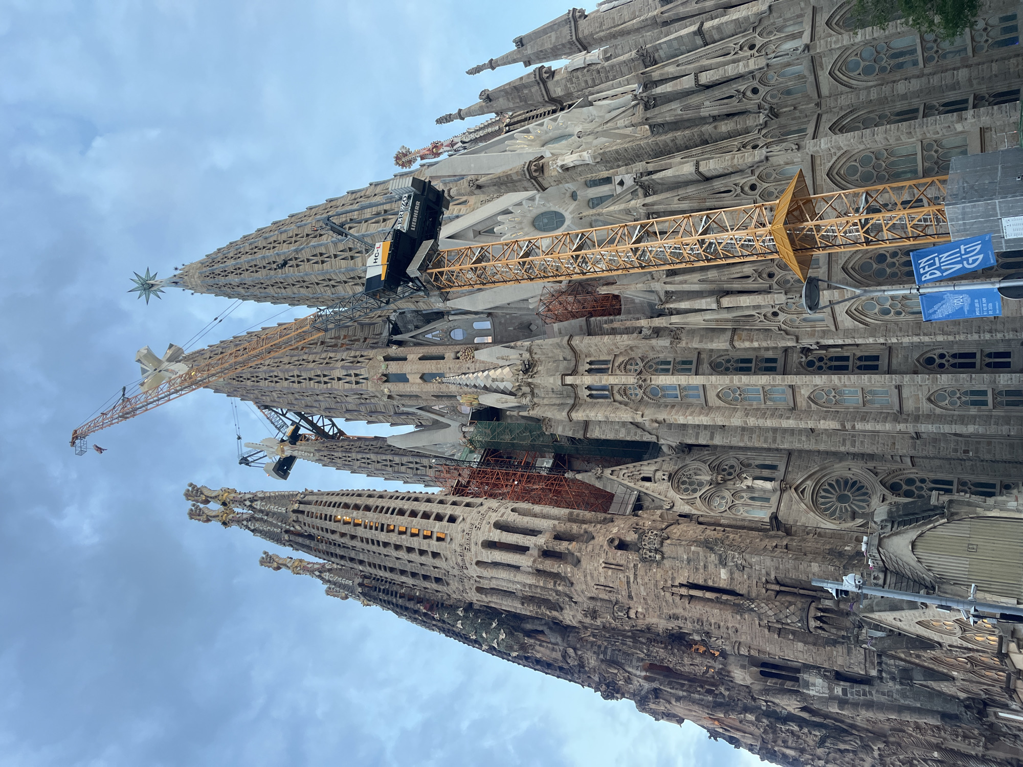 The image depicts the Sagrada Familia, a Roman Catholic basilica in Barcelona, Spain, viewed from a low angle, emphasizing its towering spires and intricate facade. The basilica is a UNESCO World Heritage site and is still under construction, as evidenced by the presence of multiple cranes. One prominent yellow crane extends from the lower right of the frame, reaching towards the central spires. Scaffolding and netting are visible on some parts of the structure, indicating ongoing work.
The architecture is characterized by its Gothic Revival style, with numerous spires adorned with ornate carvings and sculptures. The facade is made of stone, with detailed stonework and many windows. The sky above is cloudy and overcast, casting a diffused light on the scene. In the foreground, to the left, a lamppost with two blue banners is visible. The banners contain text, with one clearly showing "BEN VIN". The overall impression is one of grandeur and ongoing development of an iconic architectural masterpiece.