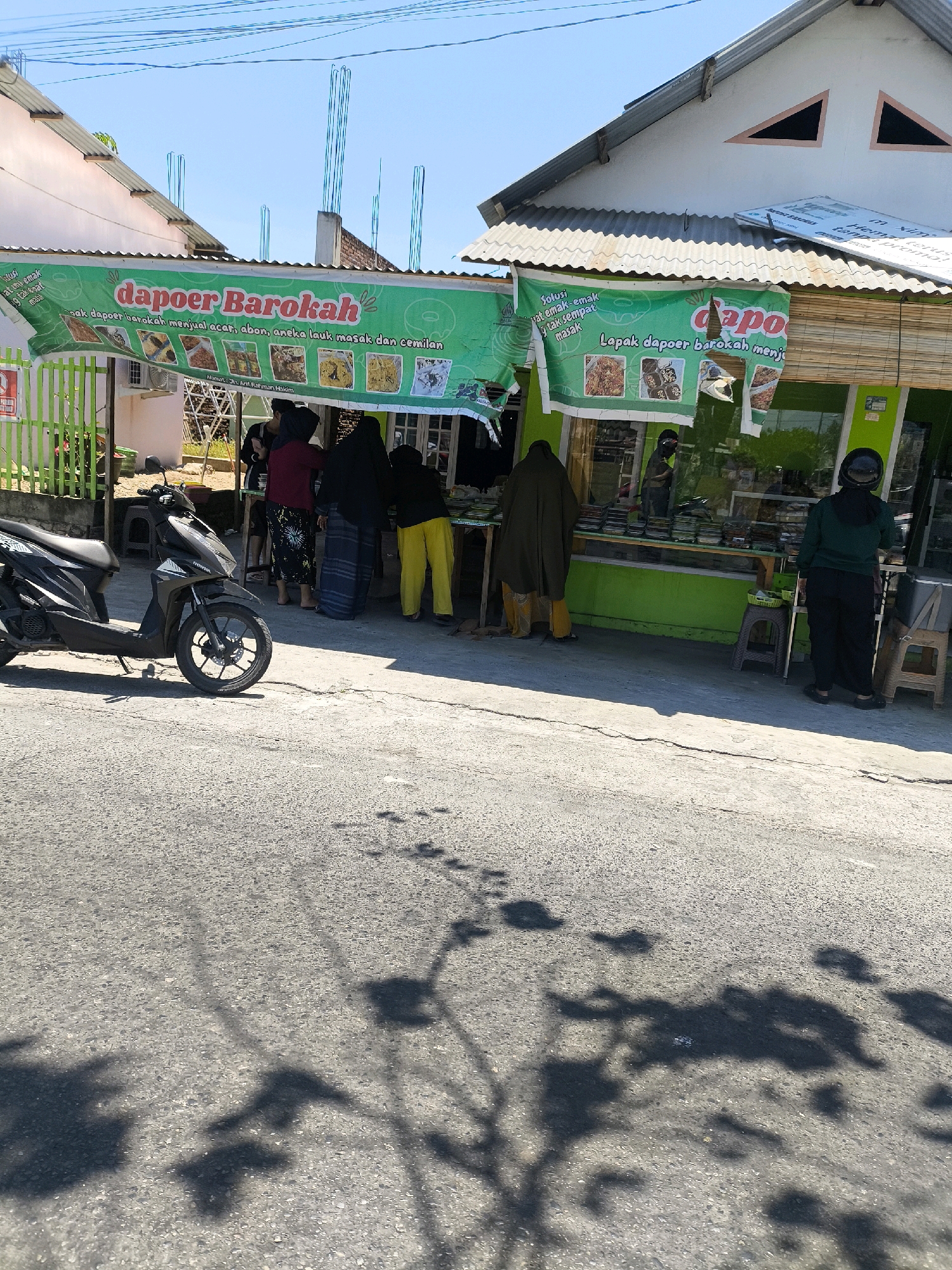 A street-level, eye-level, medium shot captures a food stall named "dapoer Barokah" in Gorontalo, Indonesia. The stall features a green awning with images of food and text in Indonesian, indicating it sells various cooked meals and snacks. Several people, some wearing headscarves and traditional Indonesian clothing like sarongs, are gathered around the stall, possibly customers or vendors. A black Honda scooter is parked on the left side of the image. The stall is situated along a paved road, with shadows from trees cast on the ground. The background shows buildings under construction, with visible rebar protruding from concrete structures, suggesting ongoing development in the area. The sky is clear and blue.