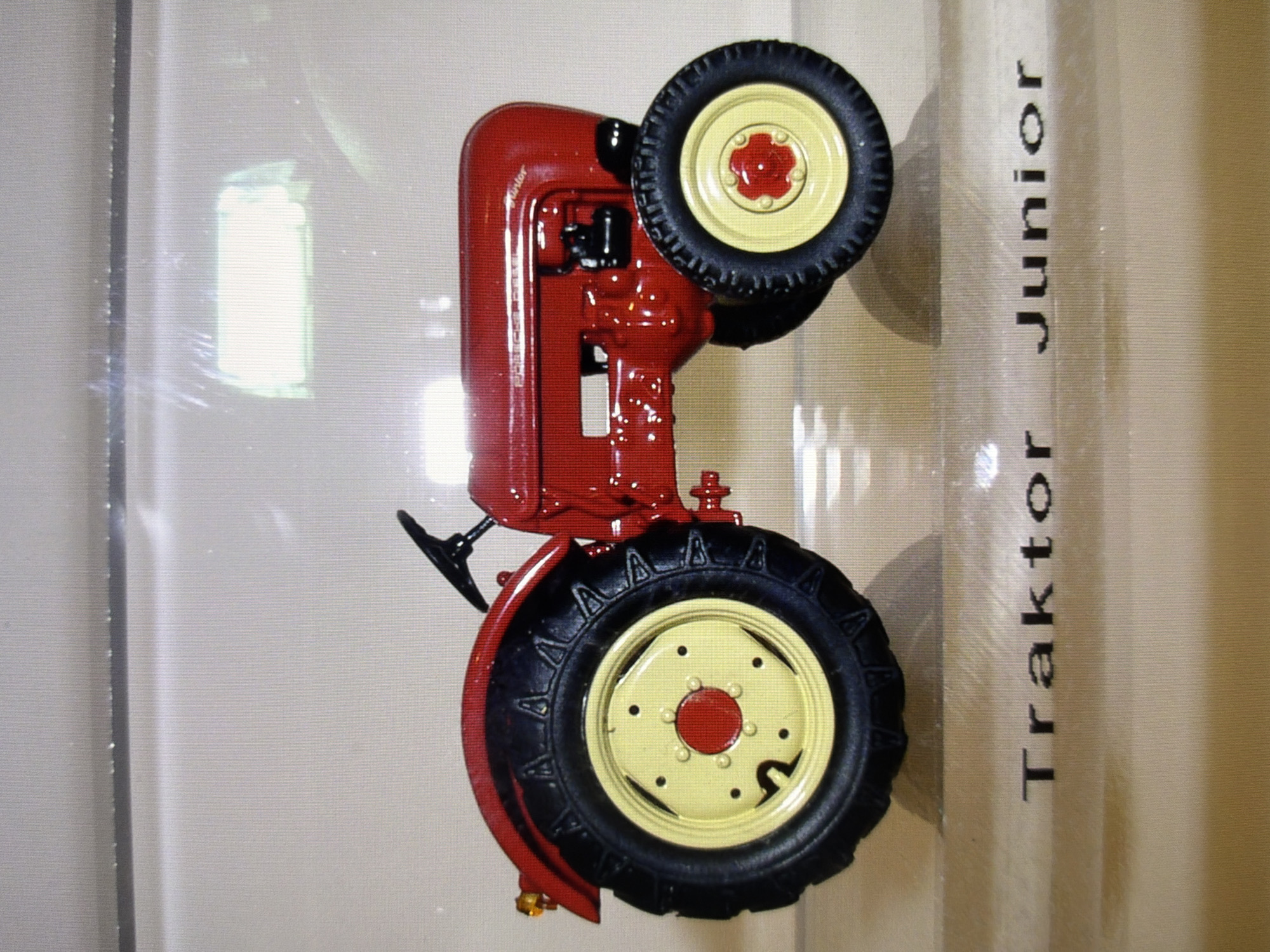 A red toy tractor, identified by the text "Traktor Junior" printed below it, is the central subject of the image. The tractor is depicted in profile, facing left, and features yellow rims on its black tires, a red body, and a black steering wheel. A small orange light is visible on the left fender. The background appears to be a light-colored, possibly textured surface, with some reflective elements, suggesting it might be displayed within a case or on a shelf. The text "Traktor Junior" is clearly legible, rendered in a dark, serif font. The image does not contain any elements that would suggest a specific location in Kyiv, Ukraine.