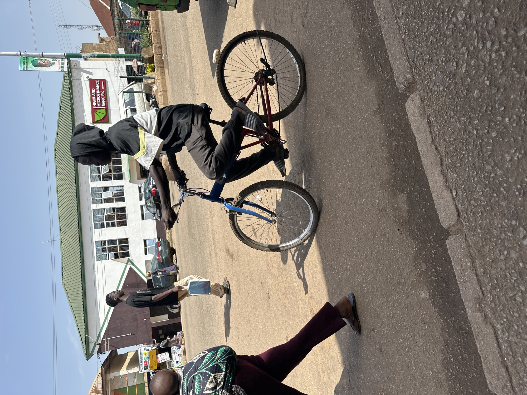 The image depicts a street scene in Bukuru, Nigeria, under a clear sky. In the foreground, a person rides a bicycle with a blue frame and red accents. The rider wears dark pants and a hooded shirt with a black, white, and yellow design. To the left, a person walks away from the camera, wearing dark purple pants and a patterned green and black top. A blue bag is visible on the ground near a person walking in the background, who is dressed in a black t-shirt and light-colored pants. A building with a green roof and white and brown facade, identified by a sign as "HIGHLAND MICROFINANCE BANK PLC", is situated behind the cyclist. A banner with a portrait of a man and the text "MUTEWANG 4+4 CONTINUITY 2027" hangs from a pole on the right side of the building. The street is paved with asphalt, and a concrete curb is visible in the lower right corner. Other vehicles and buildings are present in the background.