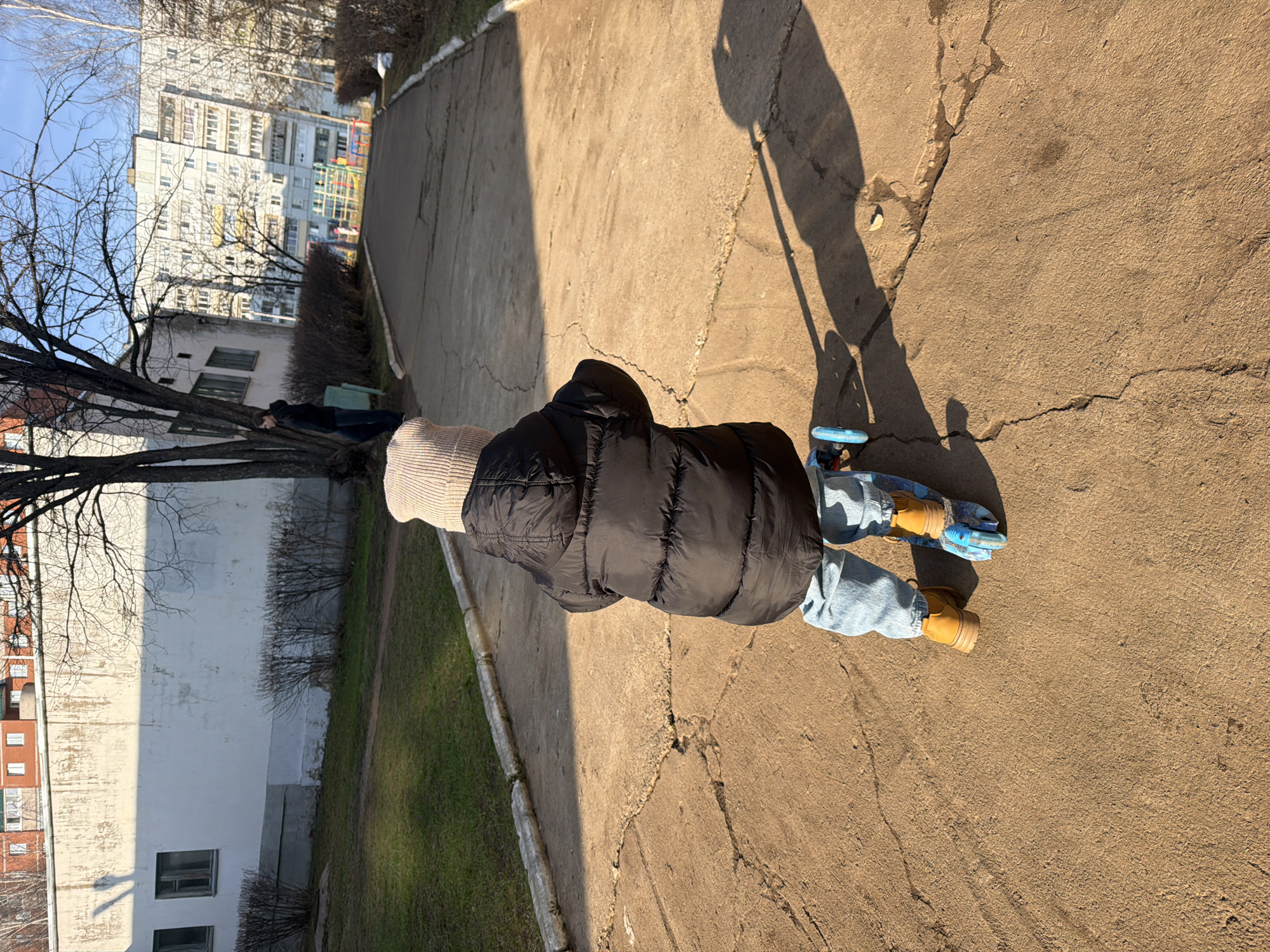 A high-angle shot shows a person standing on a concrete surface in an urban setting, likely Chaikovsky, Russia. The person wears a black puffer vest over a light-colored sweater, blue jeans, and yellow work boots. They stand on a blue skateboard. The long shadow of the photographer falls onto the surface, casting a clear line of the legs and possibly a camera holding a stick. To the left, trees and grass verge into the concrete, and in the background, a tall residential apartment building is visible under a blue sky.