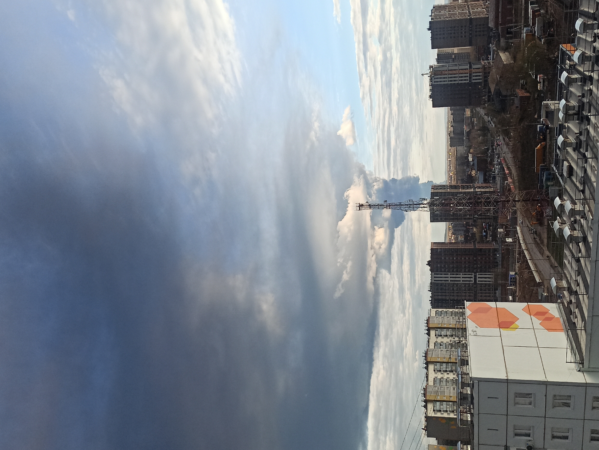 The image presents an urban landscape under a dramatic and contrasting sky, viewed from an elevated perspective. The sky is a prominent feature, divided between heavy, dark, bluish-grey clouds on the left, suggesting an approaching storm or dense weather, and brighter, more scattered cloud formations with patches of pale blue sky on the right.
The scene below the horizon is filled with numerous multi-story buildings, indicative of a modern city undergoing development. Several tall, dark-colored residential or commercial buildings dominate the mid-ground and background. Many of these appear to be under construction, with visible scaffolding and unfinished structures, while others seem completed. Towering construction cranes are scattered throughout the cityscape, with one particularly prominent crane standing near the center-right, emphasizing the ongoing building activity.
In the foreground, on the right side, an apartment building features a distinctive artistic mural on its exterior, composed of orange and yellow geometric shapes, primarily hexagons, against a white background. Adjacent to this building, another structure displays rows of rooftop or balcony-mounted air conditioning units. A glimpse of a road with vehicles can be seen amidst the buildings, confirming an active urban environment.
The time of day appears to be daytime, possibly late afternoon, influenced by the cloud cover. There are no discernible people or explicit textual elements visible in the image. The overall impression is one of a dynamic city in a state of continuous construction, set against a powerful and ever-changing sky.