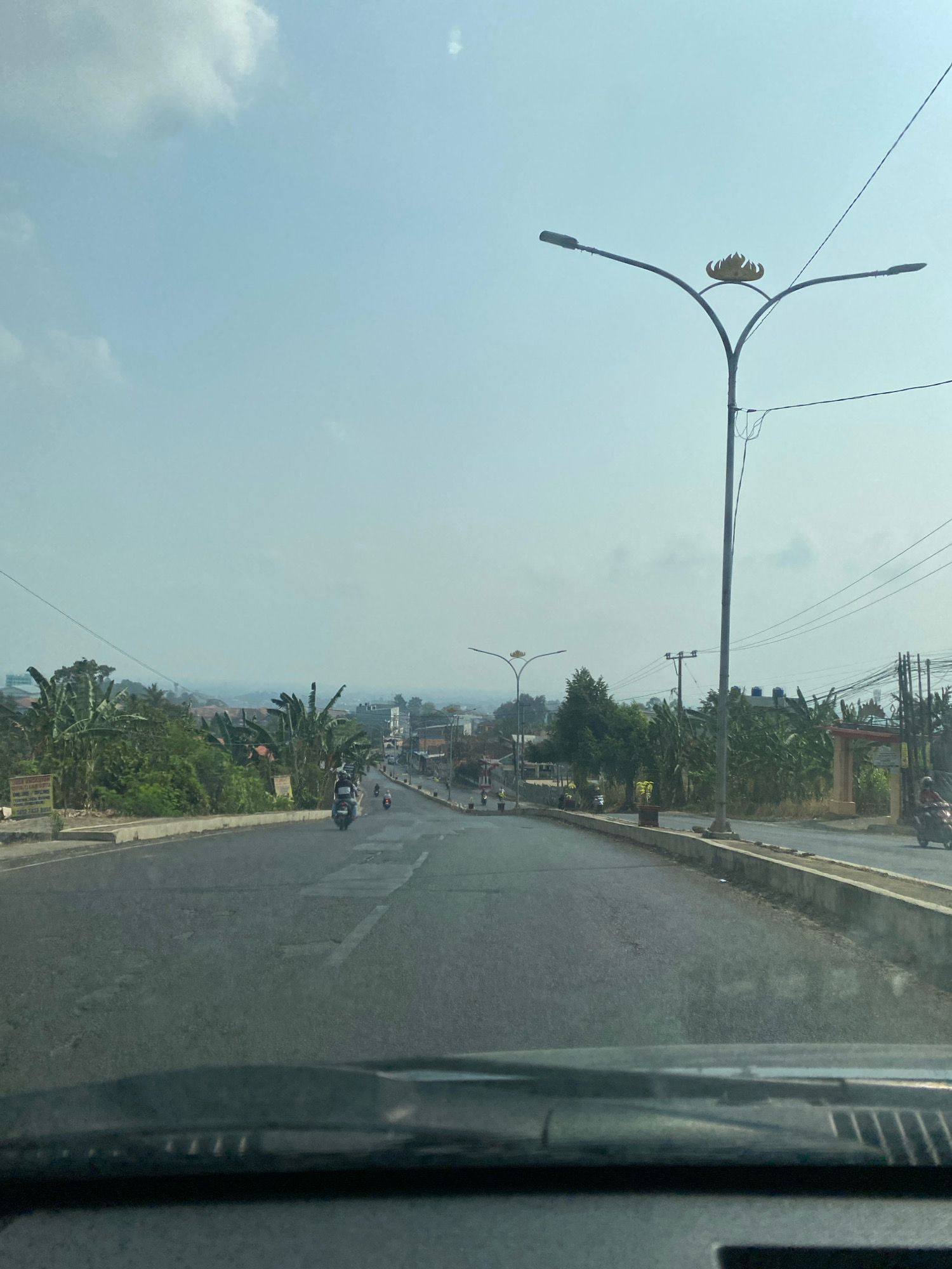 The image captures a view from a vehicle looking down a slightly uphill, paved road in Bandar Lampung, Indonesia. The road appears to have multiple lanes and is lined with a concrete barrier and vegetation on either side. Several motorbikes are visible on the road, suggesting active traffic. In the background, there are buildings and lush green trees, indicating a semi-urban or suburban environment. The sky is clear with a few scattered clouds. A prominent street lamp with an ornate, flame-like decoration is visible on the right side of the road. Power lines are also present, crisscrossing the scene. The overall impression is a sunny day in a bustling Indonesian city.