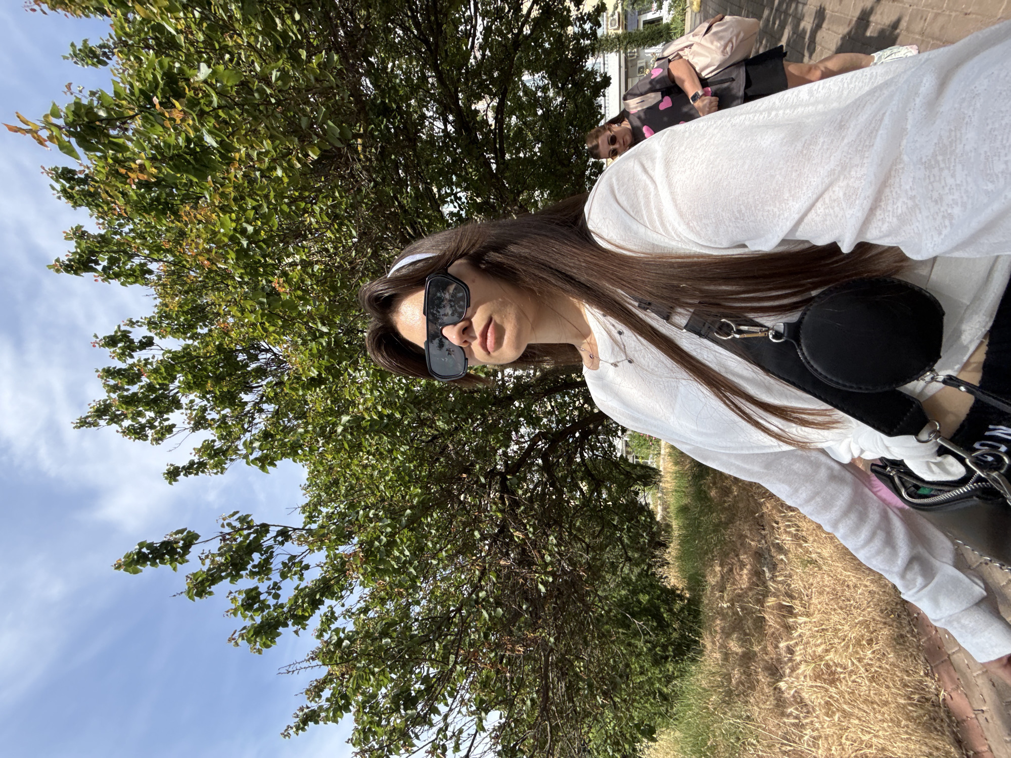 A young woman wearing sunglasses, a white long-sleeved shirt, and a white headband is standing outdoors. She has long brown hair and is looking towards her left. She is carrying a black crossbody bag. In the background, another woman wearing a black t-shirt with pink hearts and a black skirt is walking away from the camera, carrying a pink backpack. They are on a paved path next to a grassy area with a large tree. The sky is blue with scattered clouds. The scene appears to be in a park or outdoor public space.
