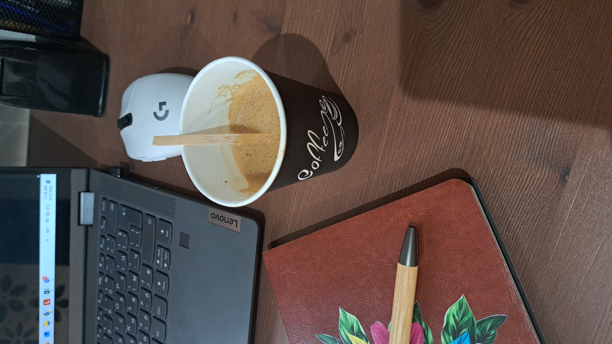 A top-down shot reveals a wooden desk with a cup of coffee positioned near a Lenovo laptop, a white computer mouse, and a notebook with a pen. The coffee cup is black with "Coffee" written on it in a whimsical white font, and a wooden stirrer is visible inside. The laptop's keyboard is partially visible, and its screen shows the Windows interface. The notebook is brown with a floral design on the cover, and a wooden pen rests on top. The setting does not provide enough information to establish a location in a specific city or region.