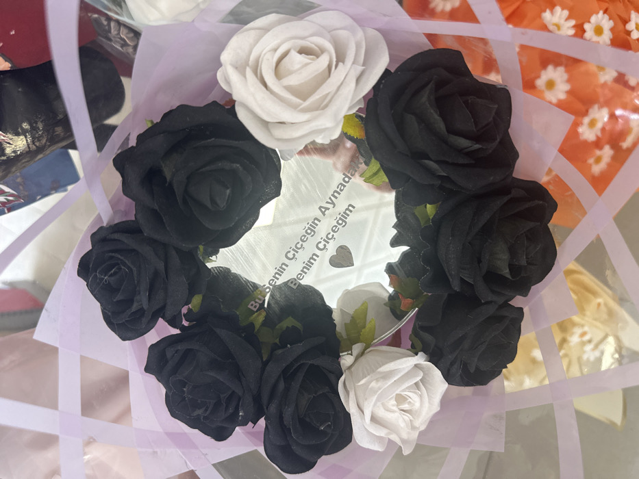 A close-up, top-down view depicts an arrangement of artificial flowers and a mirrored surface, presented in an unspecified indoor setting within the city of Torbalı, Turkey. The focal point is a circular mirror, upon which the Turkish inscription "Bu Senin Çiçeğin Aynadan Benim Çiçeğim" (translating to "This is Your Flower From the Mirror My Flower") and a small heart symbol are etched or printed. Encircling the mirror are ten artificial roses: eight black roses and two white roses, with the white roses positioned on opposite sides of the circular arrangement. The entire item is contained within clear cellophane wrapping, secured by translucent lavender ribbons arranged in a grid pattern. The background is out of focus, showing indistinct shapes and colors, including a partial view of orange paper with white daisy patterns on the lower right. No individuals are visible in the scene.