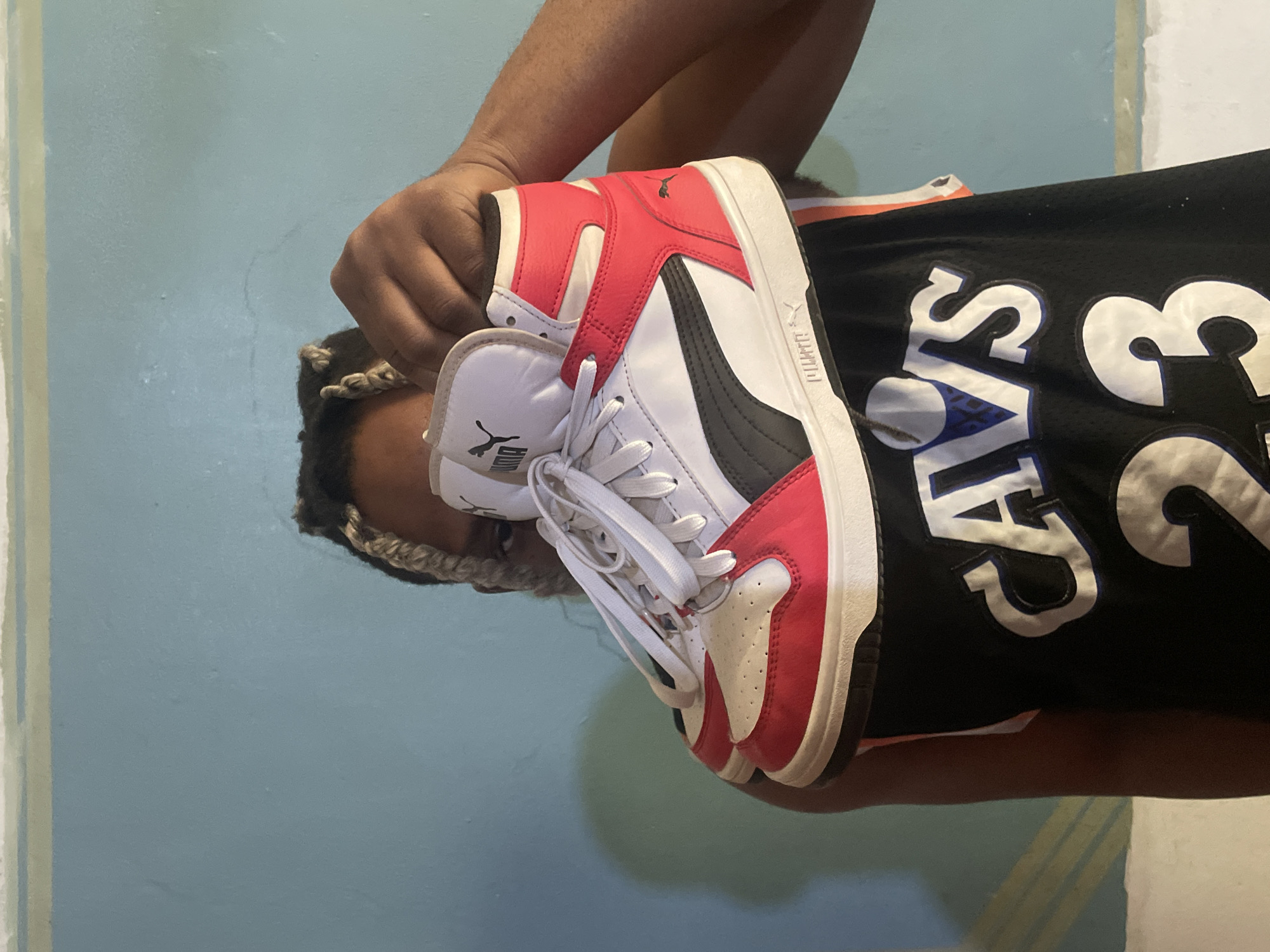 A person is holding a red, white, and black Puma high-top sneaker in front of their body. The person is wearing a black jersey with "CAVS 23" printed on it. The background is a pale blue wall. The person has dark hair with braided sections. The location is unknown.