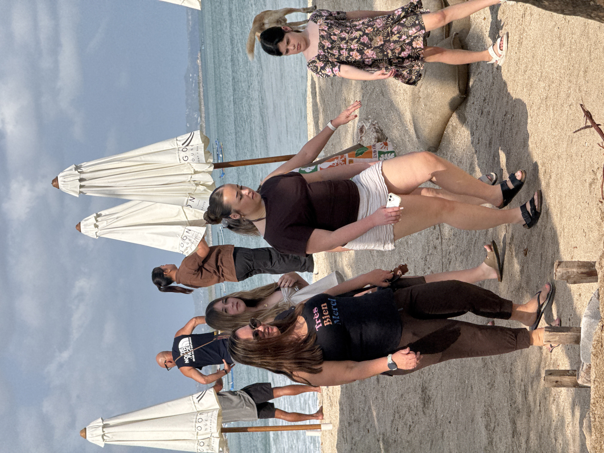 The image depicts a beach scene in Kuta, Indonesia, on a partially cloudy day. Several people are present on the sandy beach, with the ocean visible in the background. White beach umbrellas are set up, suggesting a recreational area.
In the foreground, a woman with long brown hair and sunglasses is wearing a black tank top with the text "Très Bien Merci" in pink and orange. She is also wearing brown capri pants and flip-flops. To her left, another woman with long blonde hair is wearing a light-colored top. Behind them, a man with a shaved head is wearing a black tank top with "The North Face" logo and shorts. Another woman in a brown shirt and dark pants is also visible.
Further to the right, a young woman with her hair in a ponytail is wearing a brown t-shirt and a light-colored skirt. Next to her, a young girl in a floral print dress is wearing white sandals. A dog with its tail up is walking in the background. The environment consists of sand, the ocean, and some scattered debris. Several large sandbags are visible near the right edge of the frame.
