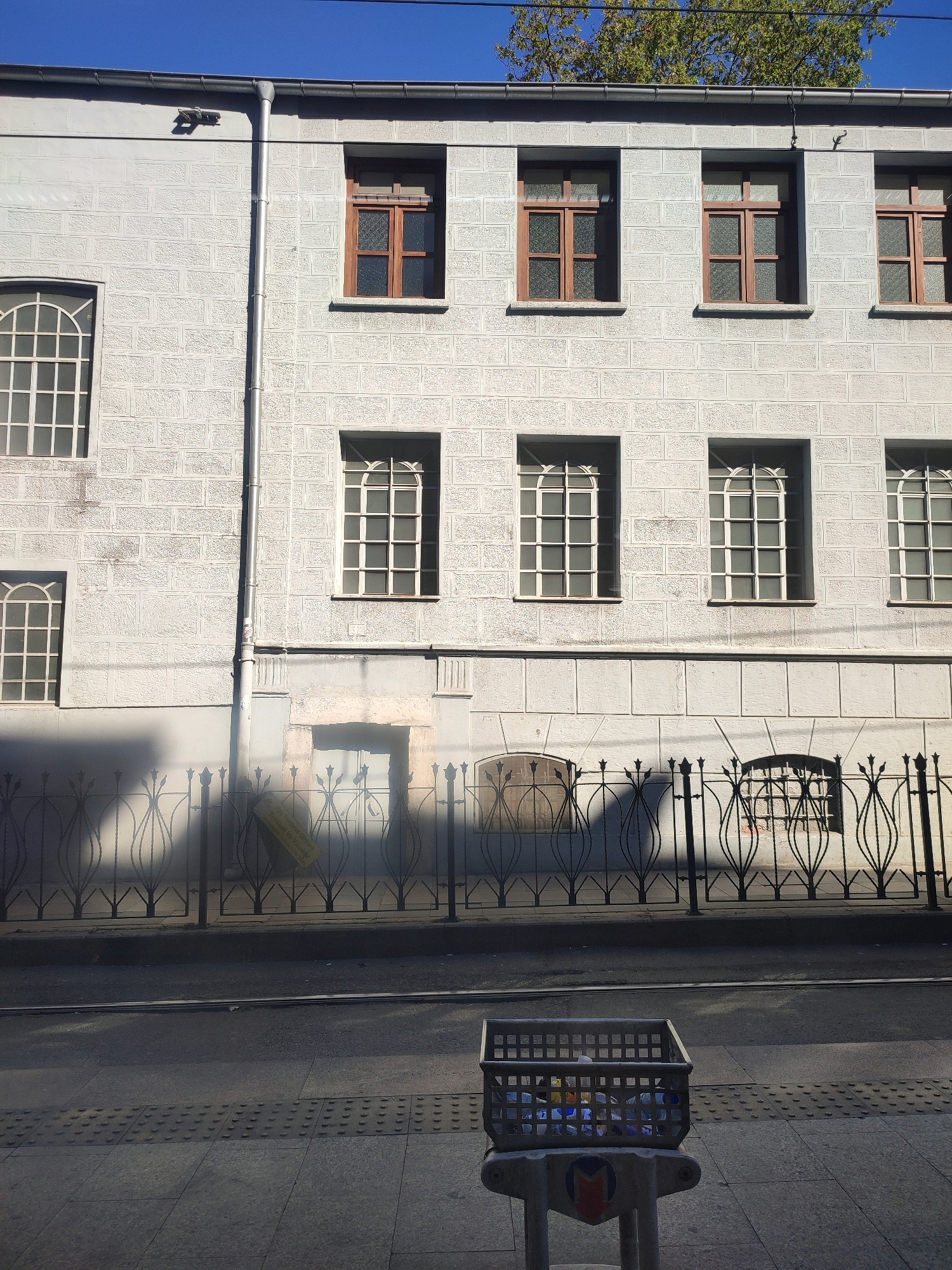 This image depicts a street view in Istanbul, Turkey, likely in the area of Beyoğlu. The scene is dominated by the facade of a light-colored building with a distinct, textured brick pattern. The building has multiple windows, some with wooden frames and patterned glass, and others with white frames and multiple panes. A downspout runs vertically along the building's exterior.
In the foreground, a black wrought-iron fence with an ornate, decorative pattern stands along the sidewalk. Behind the fence, a street is visible with tram tracks. On the sidewalk, in front of the fence, is a metal trash receptacle filled with plastic bottles and other refuse. The ground is paved with stone tiles, some of which have tactile paving for visually impaired pedestrians. Sunlight casts distinct shadows across the scene, indicating a clear, sunny day. The overall impression is one of an urban environment, possibly a historical or older district, with elements of everyday city life.