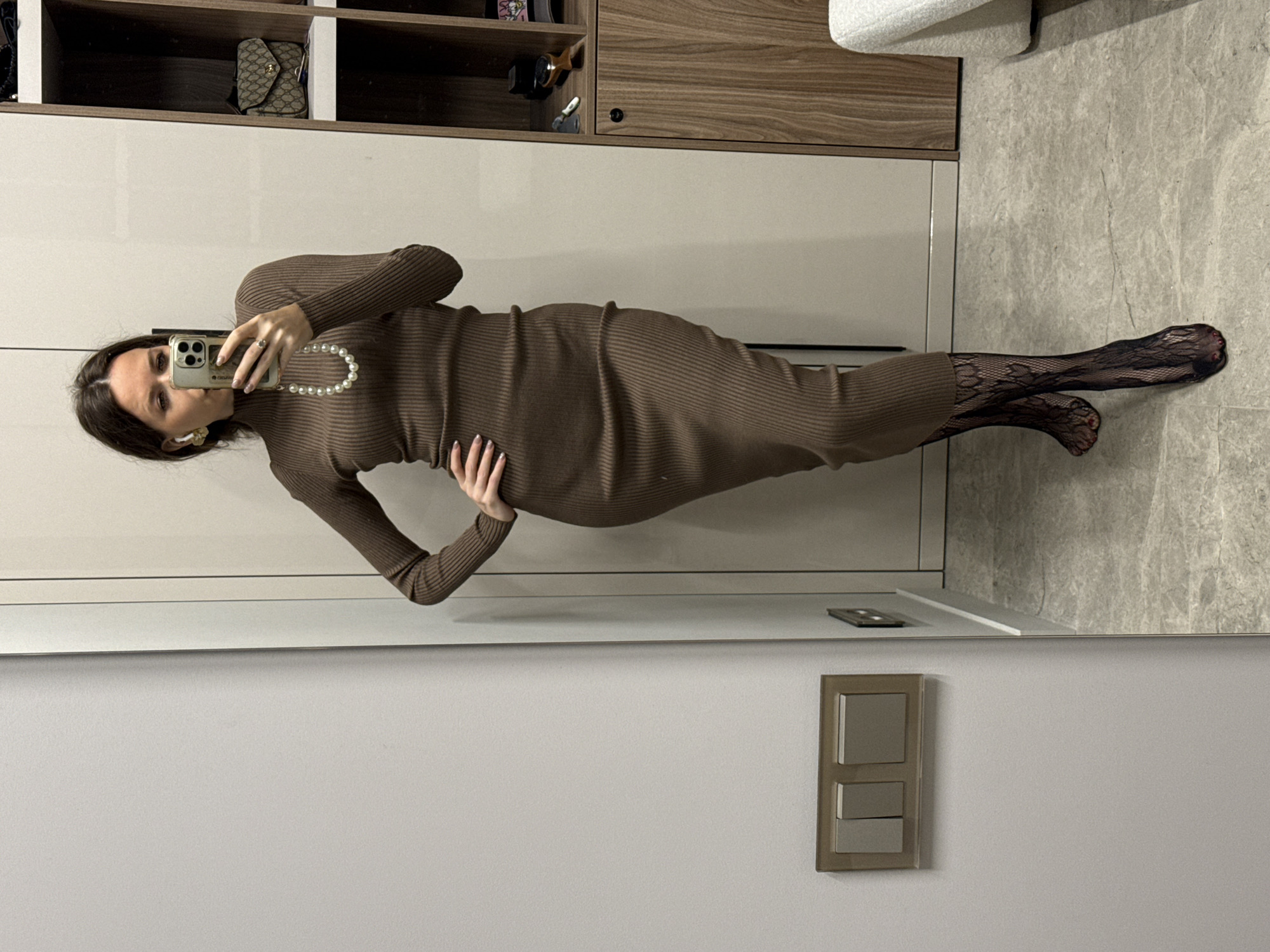 A woman stands in front of a mirror in an indoor setting. The woman is wearing a brown, long-sleeved, ribbed dress and dark patterned tights. She is also wearing a pearl necklace. She is holding a smartphone in her hand, taking a selfie. The background includes wooden cabinets and shelves, and a light-colored wall. The floor appears to be tiled. A light switch panel is visible on the wall near the bottom of the frame.