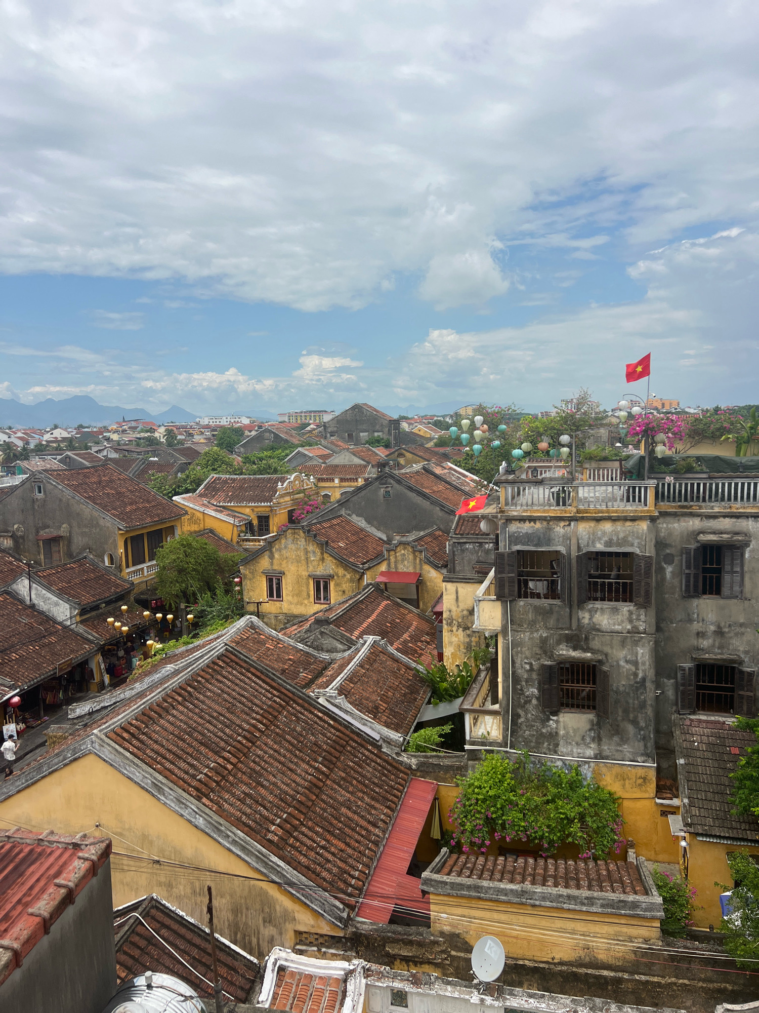 An aerial view captures the historic Hoi An Ancient Town in Vietnam. The scene is dominated by a dense cluster of traditional tiled-roof buildings, predominantly in hues of yellow and grey, with some red roofs visible. The architecture features weathered facades, wooden shutters on windows, and narrow alleyways winding between structures. Colorful lanterns are suspended along some streets, indicating a festive atmosphere. A Vietnamese flag with a yellow star on a red background flies prominently from a rooftop in the upper right quadrant of the image. In the background, distant mountains are visible under a cloudy sky. The overall impression is of a well-preserved, vibrant historical district.