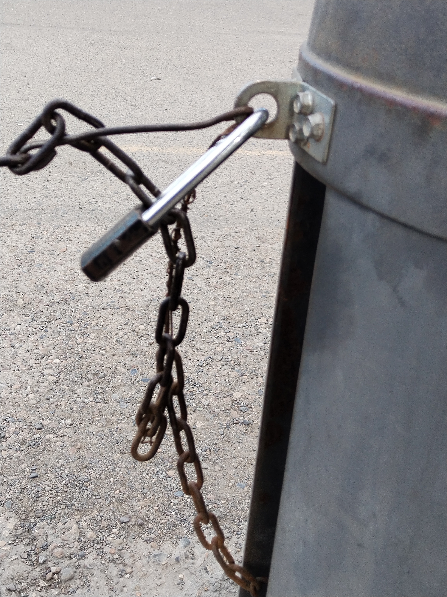 A close-up, eye-level, outdoor shot shows a metal chain and padlock securing a large, cylindrical metal object to a post. The chain is dark, appears rusty, and is looped through an eyelet attached to the cylindrical object with three bolts. A silver padlock is affixed to the chain. The cylindrical object is metallic gray and shows some signs of weathering, including a reddish-brown hue near the top. The post appears to be dark metal. The background consists of a gray asphalt surface with scattered small pebbles and dirt. The scene is static and no people are present.