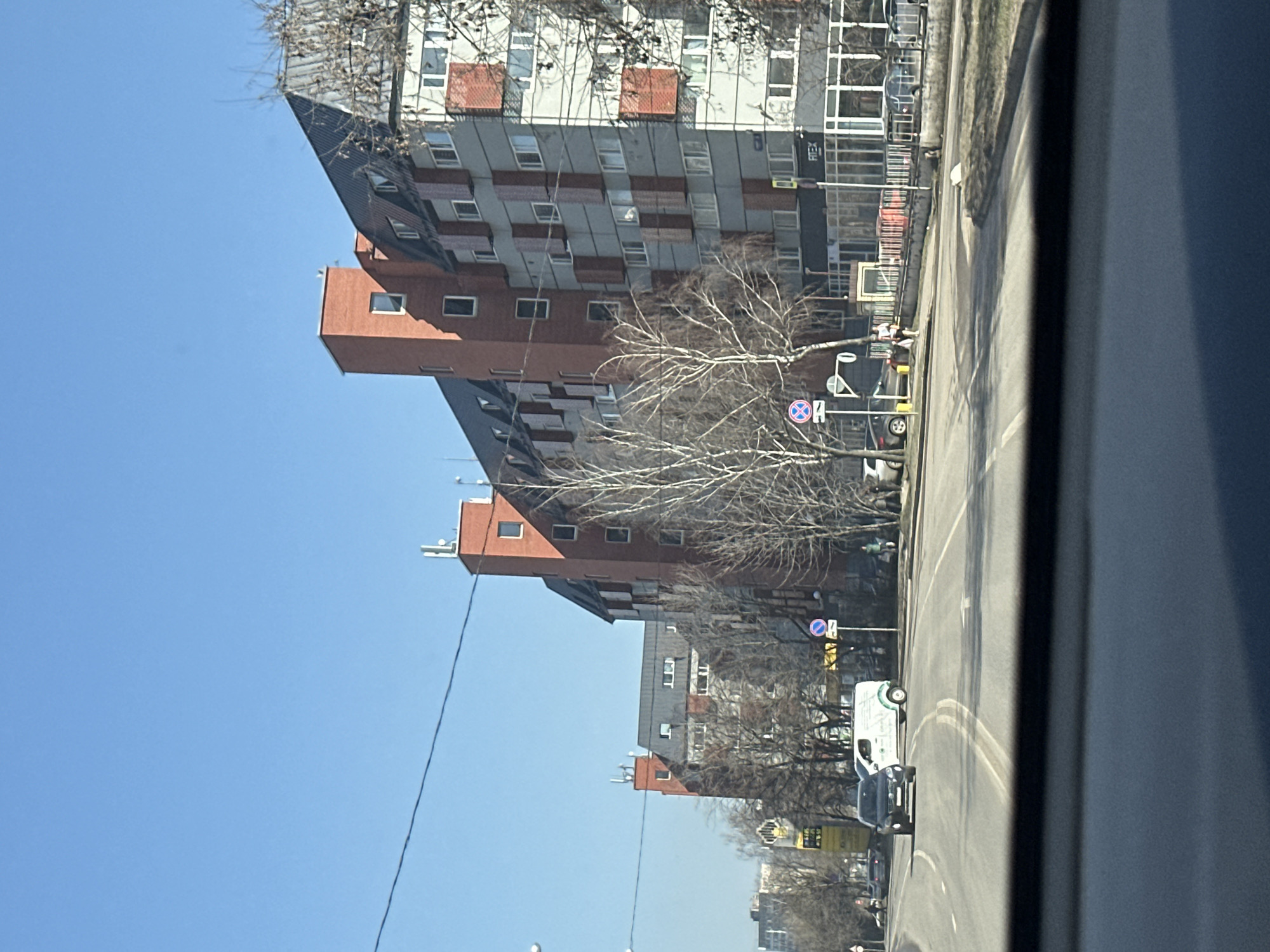 The image captures a street scene in Moscow, Russia. The view is from inside a vehicle, looking out onto a multi-lane road. A row of modern buildings lines the street, featuring a mix of red brick and gray facades with repetitive window patterns. Bare trees stand between the buildings and the road. Street signs are visible. Several vehicles are on the road, including a white van and a black car. The sky is clear and blue. The day appears sunny.