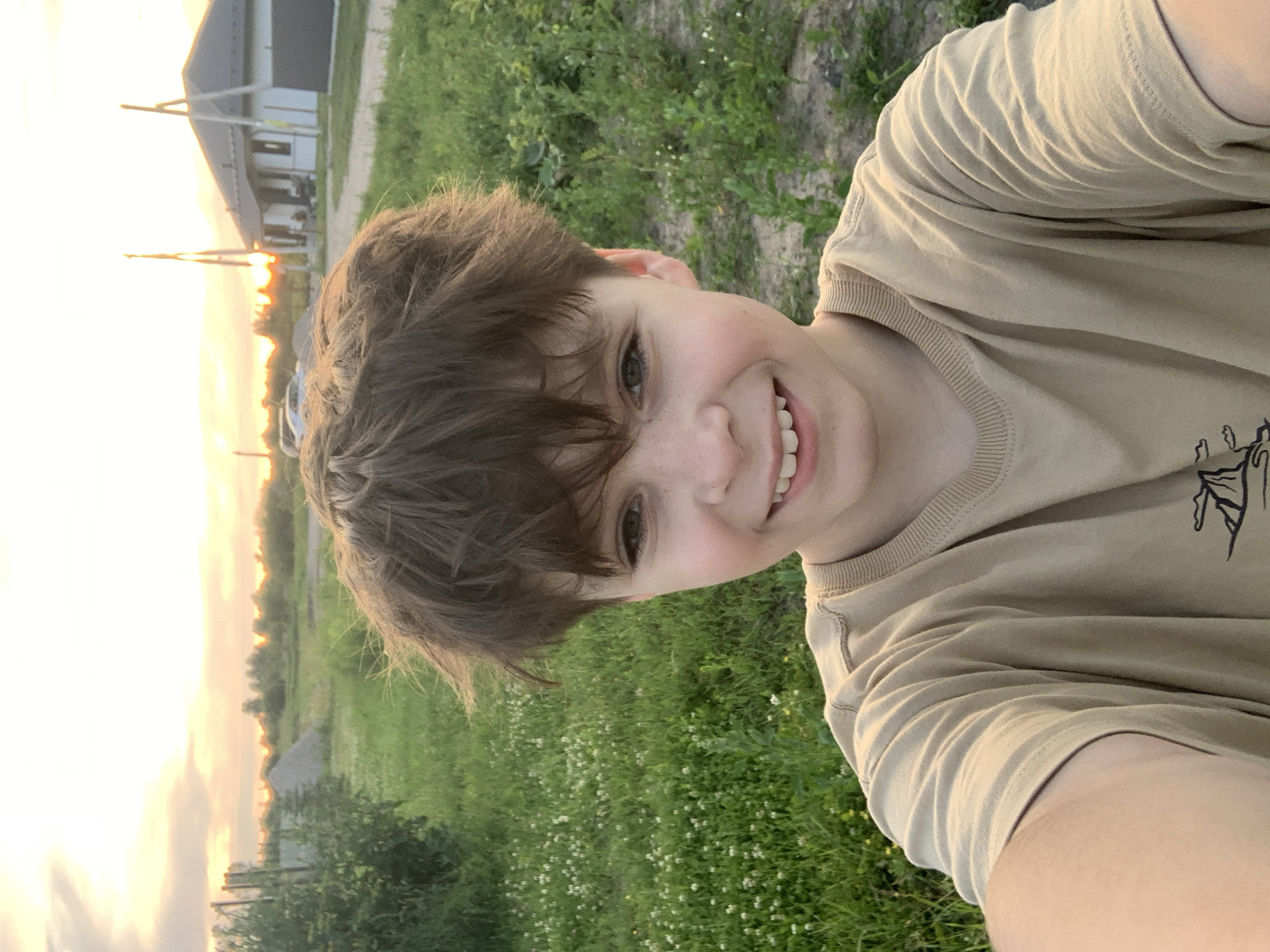 The image is a selfie taken outdoors with a subject's face and upper body prominently featured. The subject is a young person with short, tousled brown hair and a wide smile. They are wearing a light brown t-shirt. The background suggests a rural or suburban setting with green grass, some scattered white wildflowers, and a fence line. In the distance, a building with a pitched roof is visible, along with trees and a bright sky.
The lighting indicates it is likely late afternoon or early evening, with the warm glow of sunset visible on the left side of the image. The sky is bright with hues of orange and yellow, suggesting fair weather.
The image is taken in portrait orientation, with the subject's head tilted to the right. The overall mood of the photo is cheerful and relaxed, captured by the subject's broad smile. There is no visible text in the image. The location context provided is Gvardeysk, Russia.