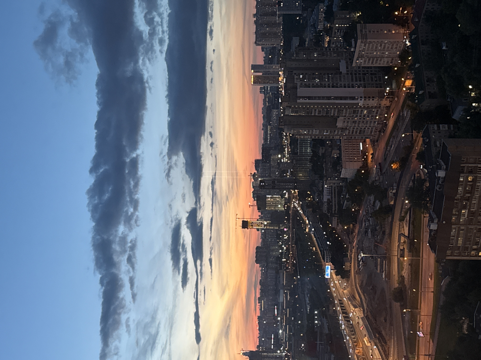 The image depicts an aerial view of a cityscape at dusk. The sky transitions from a deep blue at the top to a vibrant orange and yellow sunset near the horizon, with dark clouds scattered across the upper portion. In the foreground, there is a complex network of roads and highways with cars and trucks visible. Construction sites with cranes are interspersed with residential and commercial buildings of various heights. Many buildings are illuminated, indicating that it is after sunset. The overall scene is one of urban development and the transition from day to night.