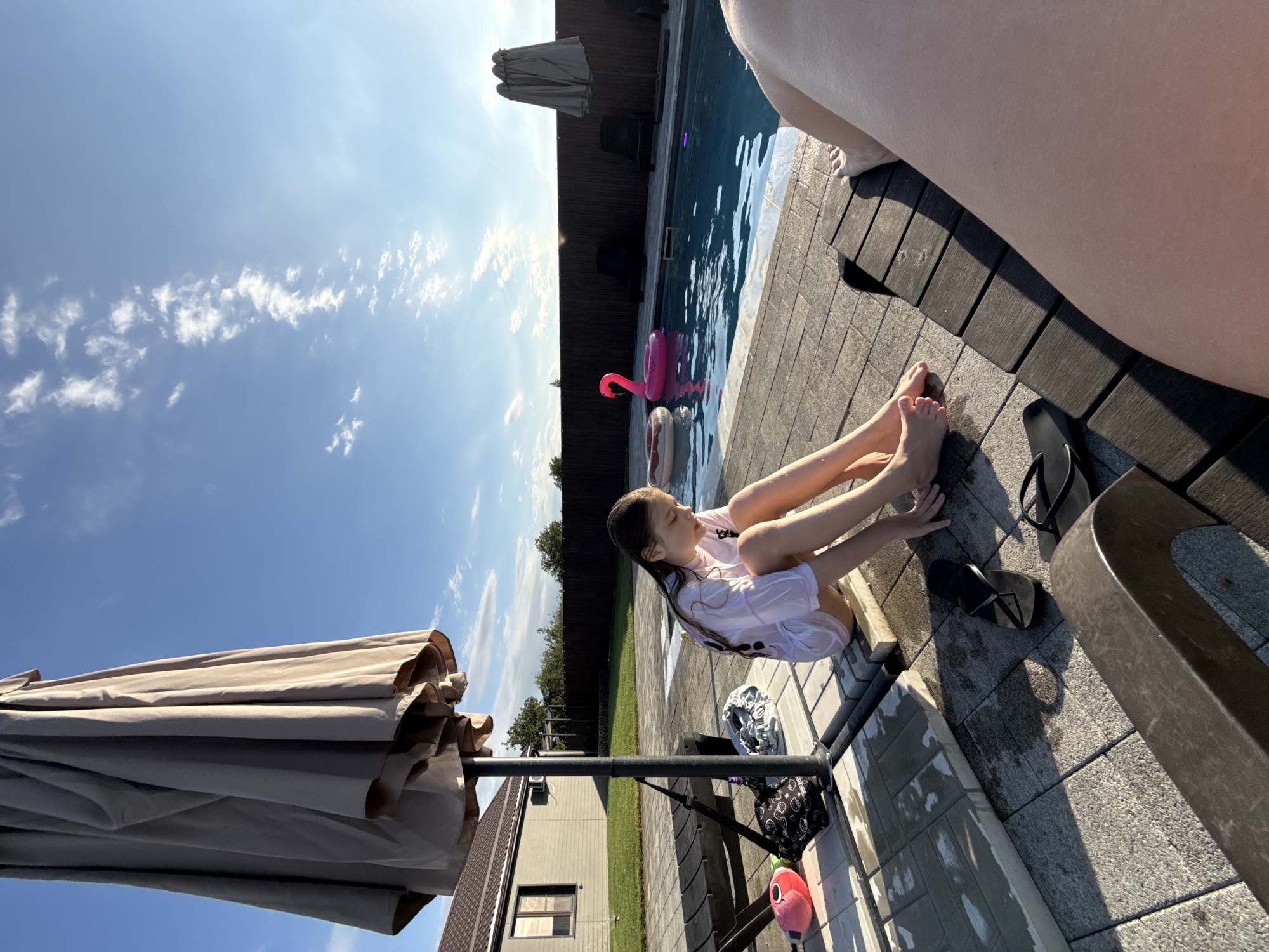 A young female with wet hair, wearing a white t-shirt and shorts, sits on the tiled edge of a swimming pool. Her feet are bare and touch the tiles. To her left, a large closed umbrella stands. Nearby are a pair of black flip-flops and a red stuffed toy. The pool contains a pink flamingo float and a donut-shaped float. In the background, a wooden fence encloses the yard. The location is Pervorechenskoye, Russia, on a sunny day with a blue sky and scattered clouds.
