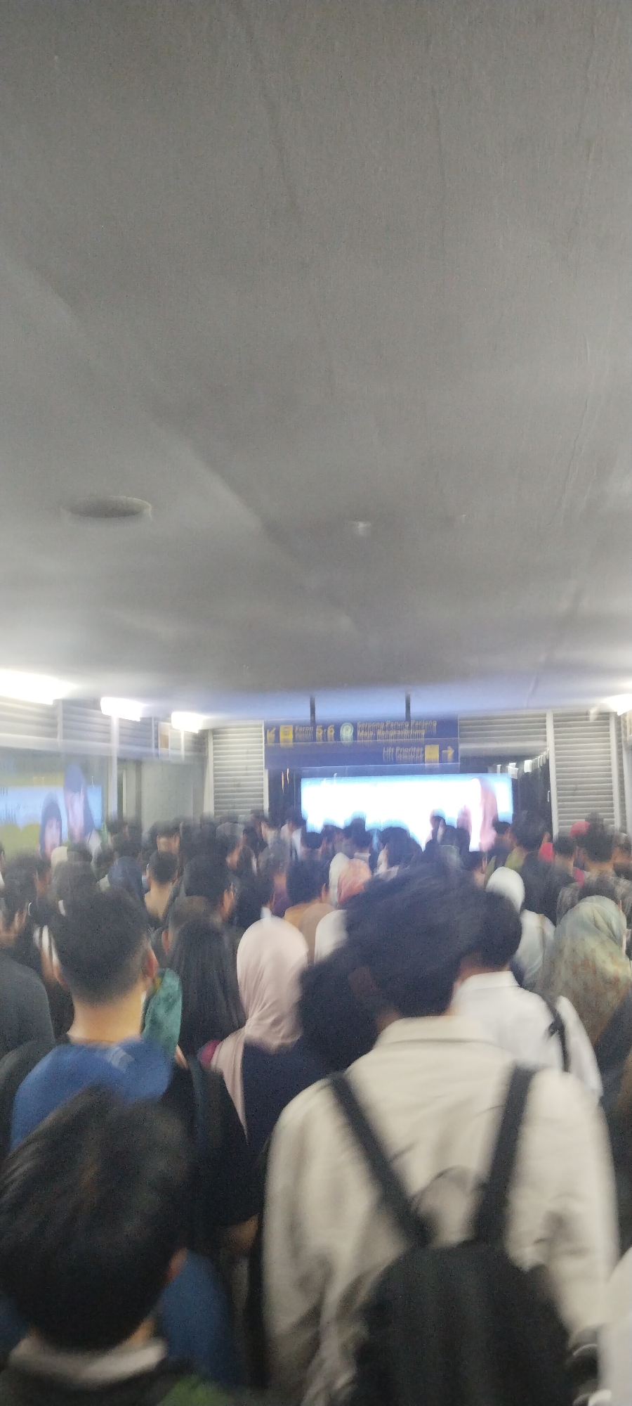 The image shows a crowded indoor scene, likely a public transportation hub or a building lobby in Central Jakarta, Indonesia. A large group of people, diverse in age and gender, are standing or moving in a densely packed area. Many are wearing casual clothing, and some women are wearing headscarves. The lighting suggests an artificial environment with overhead lights visible. In the background, a large digital screen displays text and images, possibly providing information or advertisements. Signs with Indonesian text are also visible, including one indicating "Lift Prioritas," meaning "Priority Lift." The overall atmosphere appears to be one of transit and waiting, with people navigating through the crowded space.