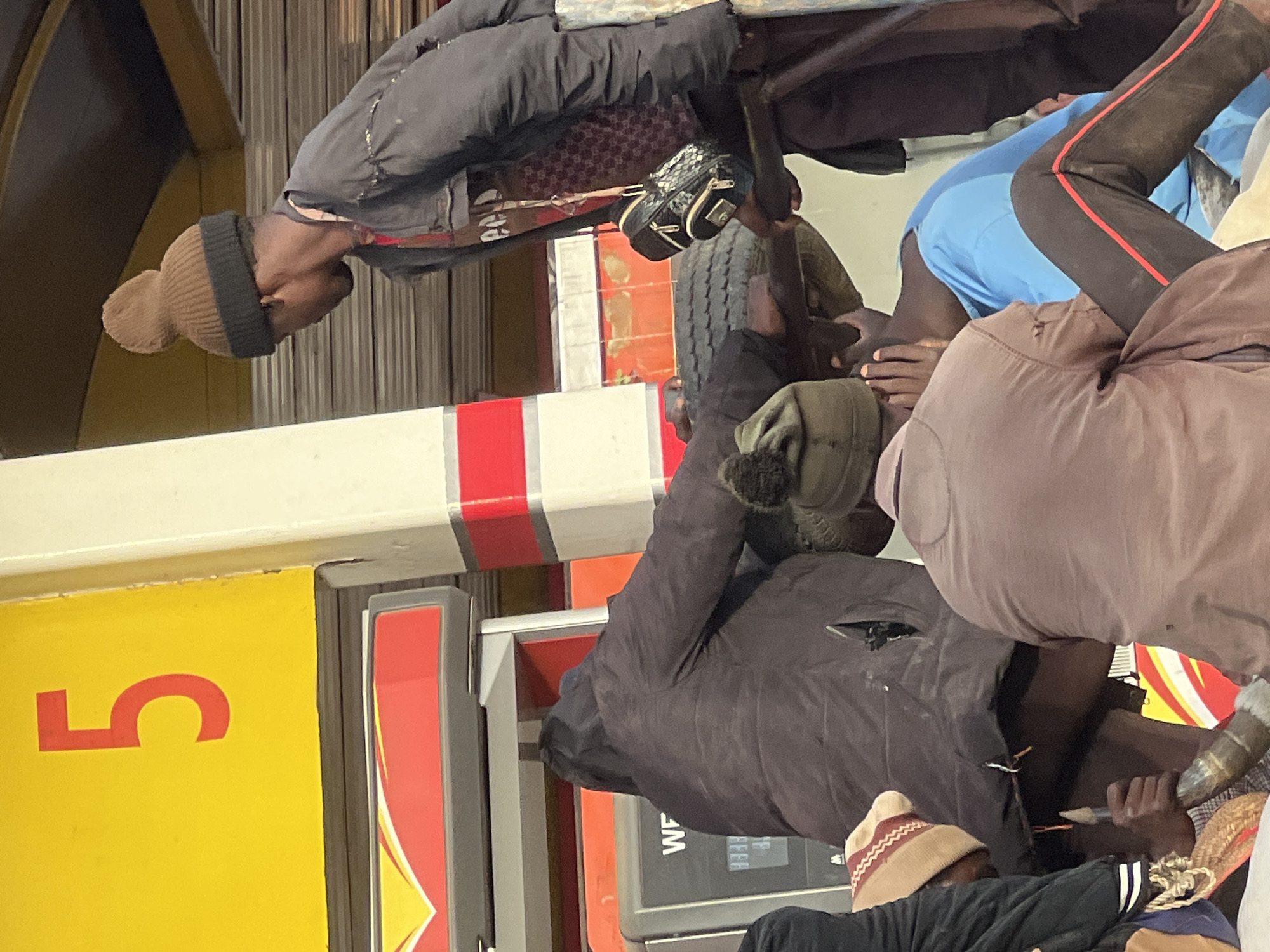 This image, taken in Jos, Nigeria, depicts several men gathered around what appears to be a fuel pump area at a gas station. The scene is likely set at night or in dimly lit conditions.
In the foreground, several men are visible. One man, wearing a brown beanie and a dark jacket, is looking down and slightly to the left. His back is to the viewer. To his right, another man in a blue shirt and dark pants with a red side stripe is partially visible, seemingly bent over or kneeling, with his hands near the ground or another person. A third man, wearing a light brown jacket, is also seen from the back. A fourth man, whose face is not visible, wears a light-colored beanie with red stripes.
Objects in the scene include a large, dark tire resting or mounted on some equipment, and what looks like the top part of a fuel pump or dispensing machine. A small, patterned bag hangs from a structure near the tire. A metal barrier with red and white stripes runs horizontally across the mid-ground.
The setting appears to be a petrol station, suggested by the characteristic yellow and red color scheme, and the prominent red number "5" on a yellow panel, which commonly denotes a pump number. On a digital display of one of the machines, the letters "WF" and some numbers, possibly "888", are faintly visible. The men seem to be waiting or resting, with no clear active event taking place, though one man's hand is near what could be a pump nozzle. Emotions are not distinctly discernible from their postures.