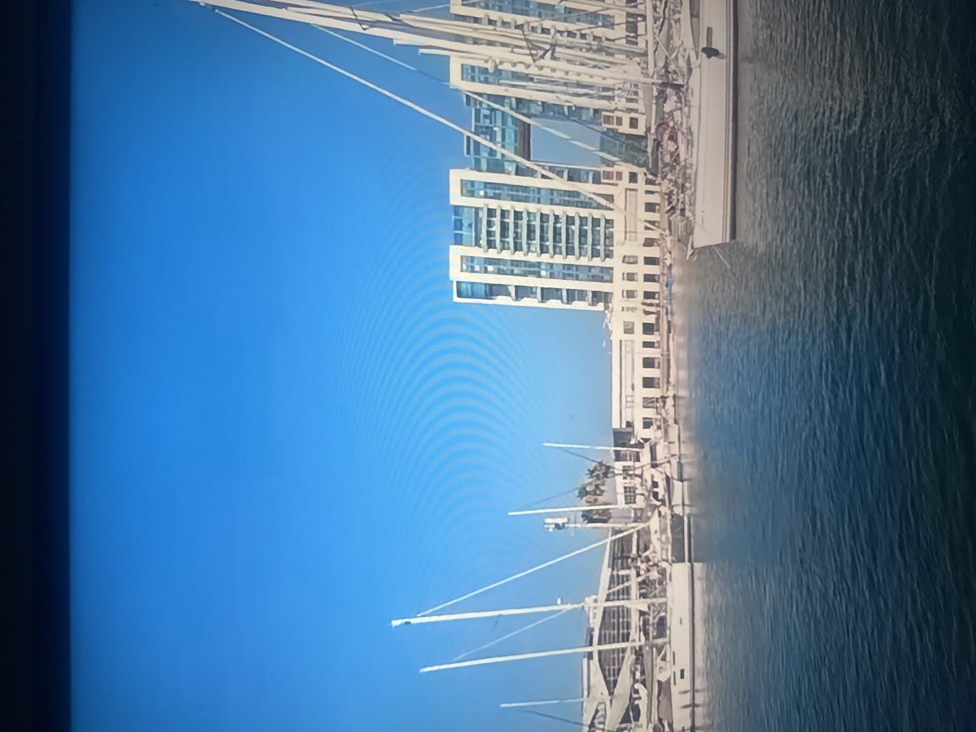 This image depicts a modern marina in Herzliya, Israel, on a clear, sunny day. The foreground is dominated by the calm, blue waters of the marina, reflecting the sky and the structures beyond. Several white sailboats are moored in the water, their masts extending upwards.
In the mid-ground, a row of buildings lines the waterfront. These buildings appear to be a mix of modern architecture, including a distinctive multi-story structure with large windows and an archway connecting two sections. Some of these buildings seem to house businesses or residences. Palm trees are visible between the buildings, adding a touch of tropical greenery to the scene.
The background is a vast expanse of clear blue sky, with faint concentric rings possibly indicating some atmospheric phenomenon or lens artifact. The overall impression is one of a well-developed urban waterfront with recreational and residential amenities. No people are clearly visible in the image. The scene is static, with the boats and buildings appearing stationary.