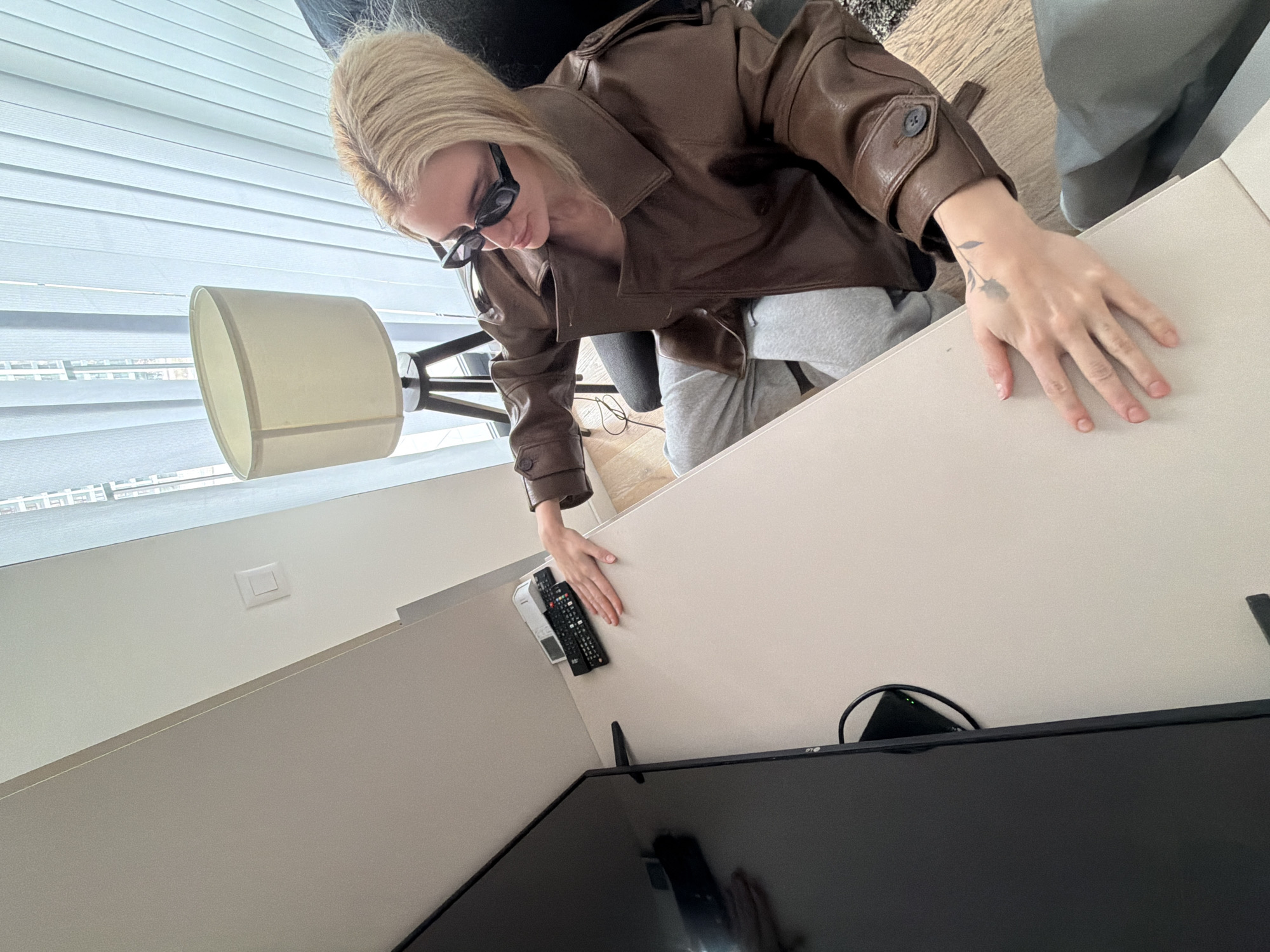Belgrade, Serbia. An indoor scene depicts a blonde female seated in a dark office chair at a light-colored desk. She is wearing a brown leather-like jacket, grey sweatpants, and black rectangular sunglasses. Her left hand, featuring a dark floral tattoo on its dorsal surface, rests flat on the desk. Her right hand is extended towards a stack of three remote controls (two black, one white) situated on a narrow shelf behind the desk. A large, black LG flat-screen television is positioned vertically on the left side of the frame, partially obscuring the desk. In the background, a floor lamp with a beige fabric shade and a dark tripod base stands near a window fitted with vertical blinds, revealing tall urban buildings outside. The room features light-colored walls and light wooden flooring, with a fragment of a dark rug visible under the chair. A white square light switch is visible on the right wall. The scene is illuminated by natural light.