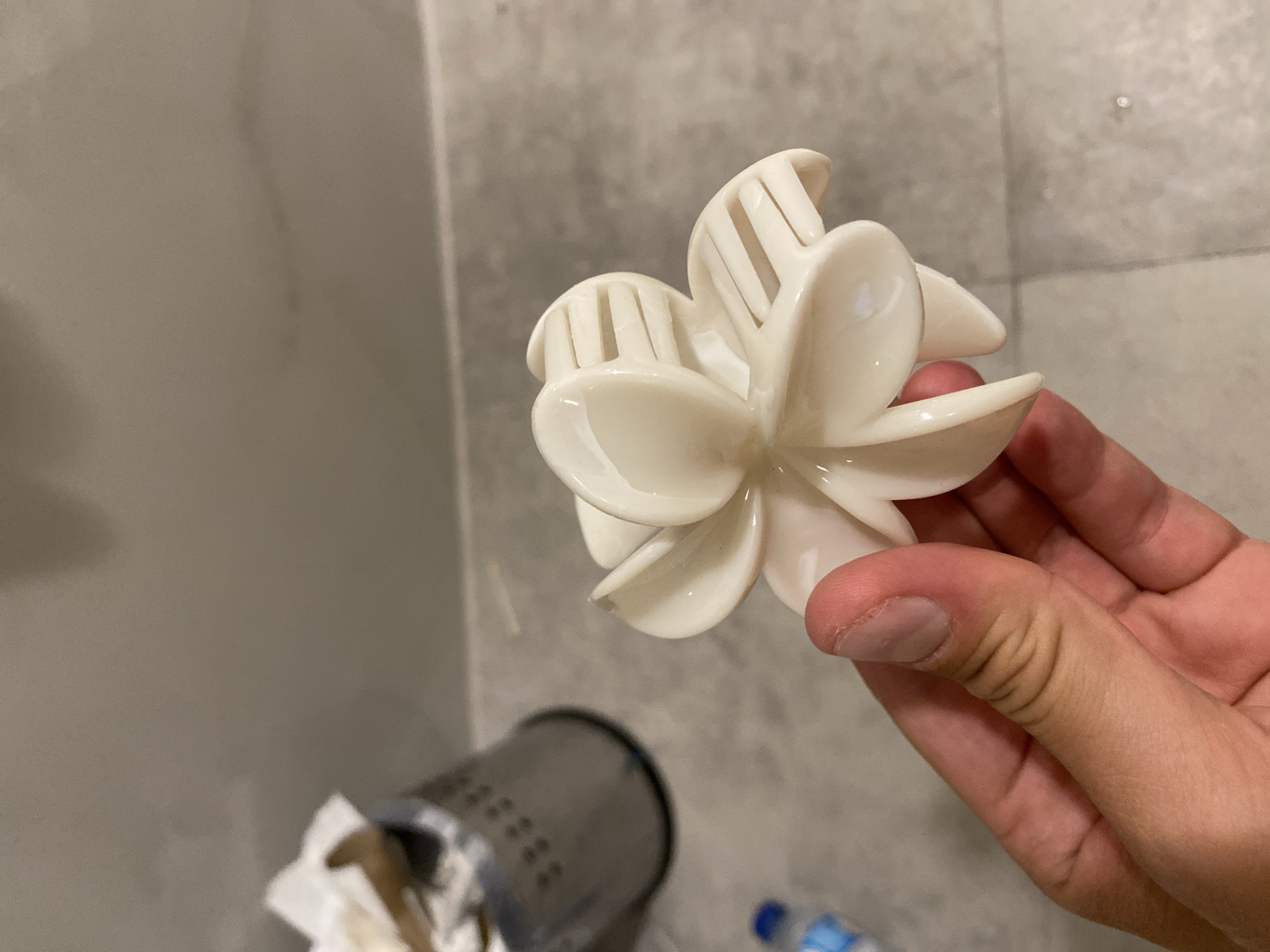 A close-up shot shows a hand holding a translucent white flower-shaped object, possibly a hair clip or decorative item. The object has six petal-like extensions, with the upper ones featuring parallel slits. The hand, with visible fingernails and skin texture, grips the object from below, partially obscuring its base. In the blurred background, a silver-colored bin with a perforated pattern is visible, containing crumpled white paper. A blue-capped bottle rests on a light-gray, tiled surface, which also appears to be the floor. The overall setting suggests an indoor environment, possibly a bathroom or a public restroom. There is no information in the image that would allow for its location to be identified as Aksu, Turkey.