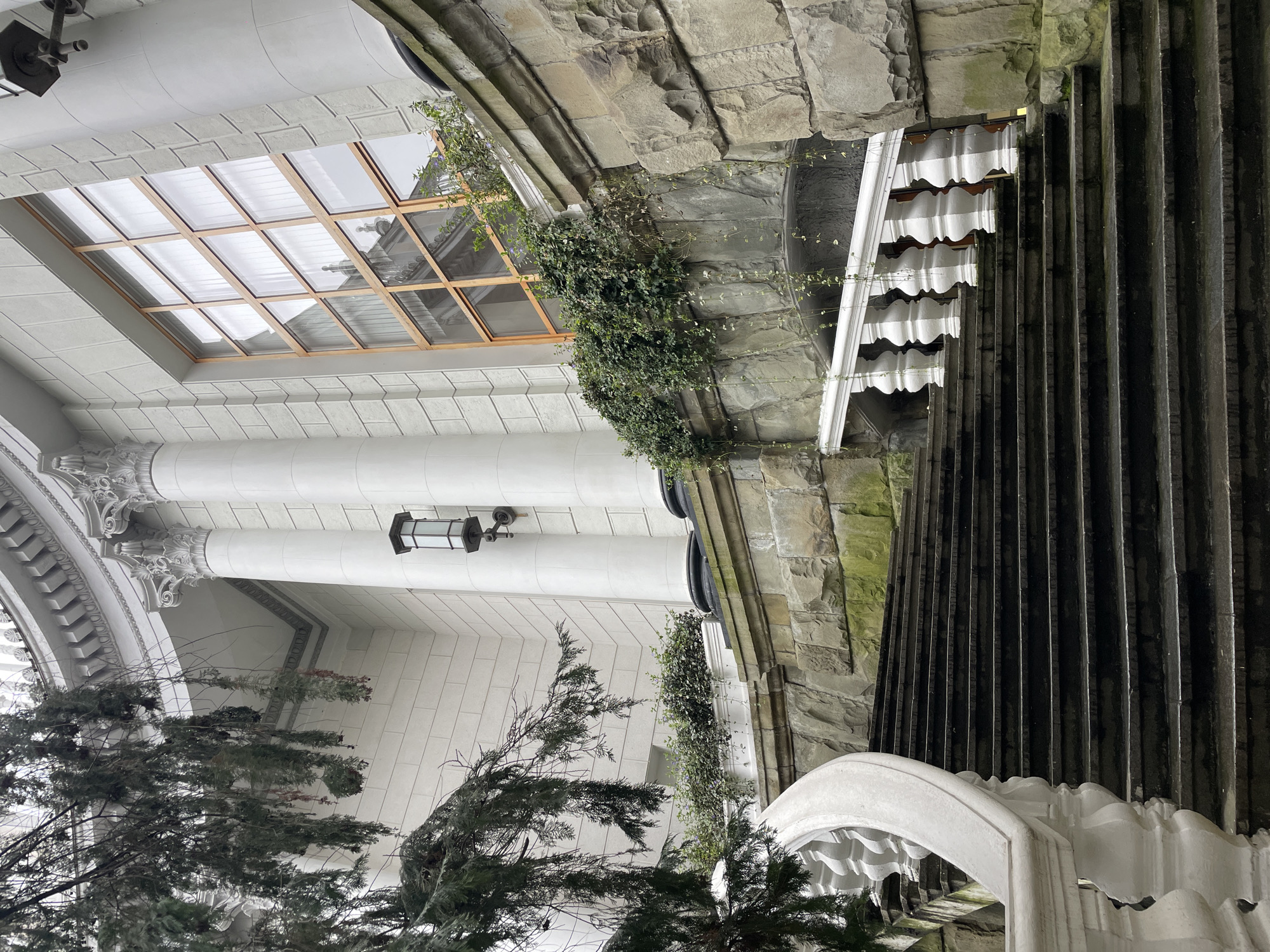 The image depicts an architectural scene, possibly an exterior view of a building in Sochi, Russia. The architecture exhibits elements of both modern and classical styles. A prominent feature is a rectangular skylight or window composed of multiple glass panes.
The building's façade is predominantly white with columns and ornate detailing. There's a wall constructed from stacked stones and overgrown with greenery.
A set of dark-colored vertical structures with a repetitive pattern (perhaps ribs or slats) ascends or descends along the right side of the image. A decorative white element with arches is visible. Evergreen trees grow in front of the structure.
There are no people visible in the image. The scene appears to be either dimly lit or the photograph was taken during twilight hours.