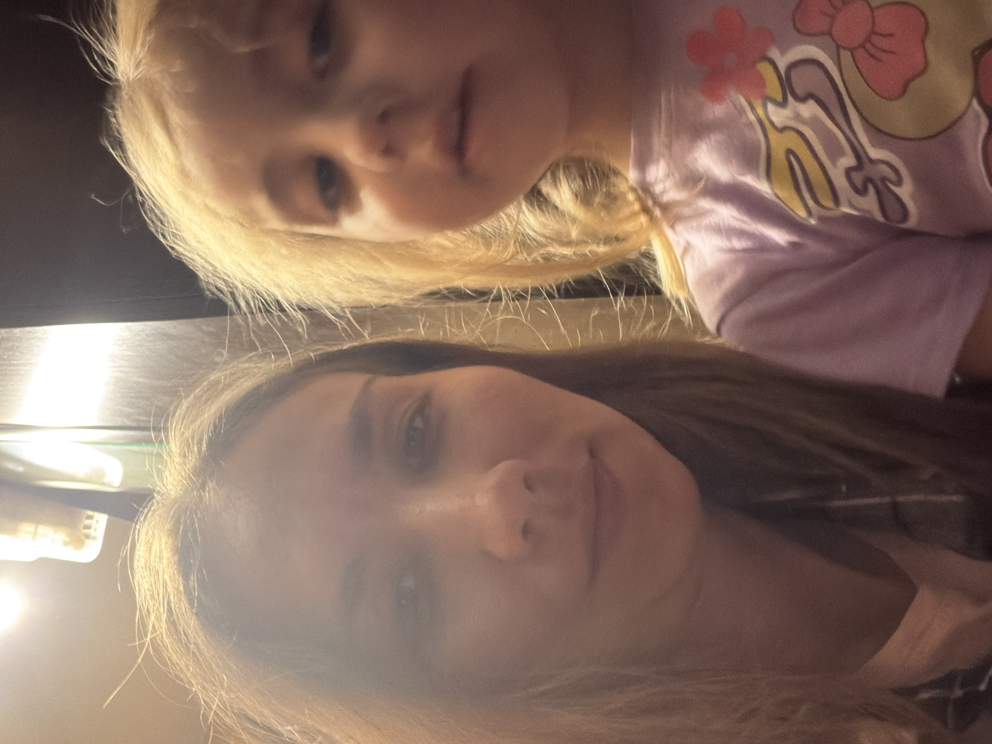 The image shows a selfie of two people, an adult female and a young child, likely a girl. The adult, on the left, has shoulder-length brown hair and is looking towards the camera. The child, on the right, has blonde hair and is also looking towards the camera, though her face is partially obscured and appears slightly out of focus. She is wearing a purple t-shirt with a cartoonish design featuring flowers and what appears to be a character. The background is dimly lit, with a bright light source visible in the upper left corner, suggesting an indoor setting. The overall scene is casual and personal, typical of a selfie. There are no discernible landmarks or features that would indicate the specific location within the city of Tambov, Russia.