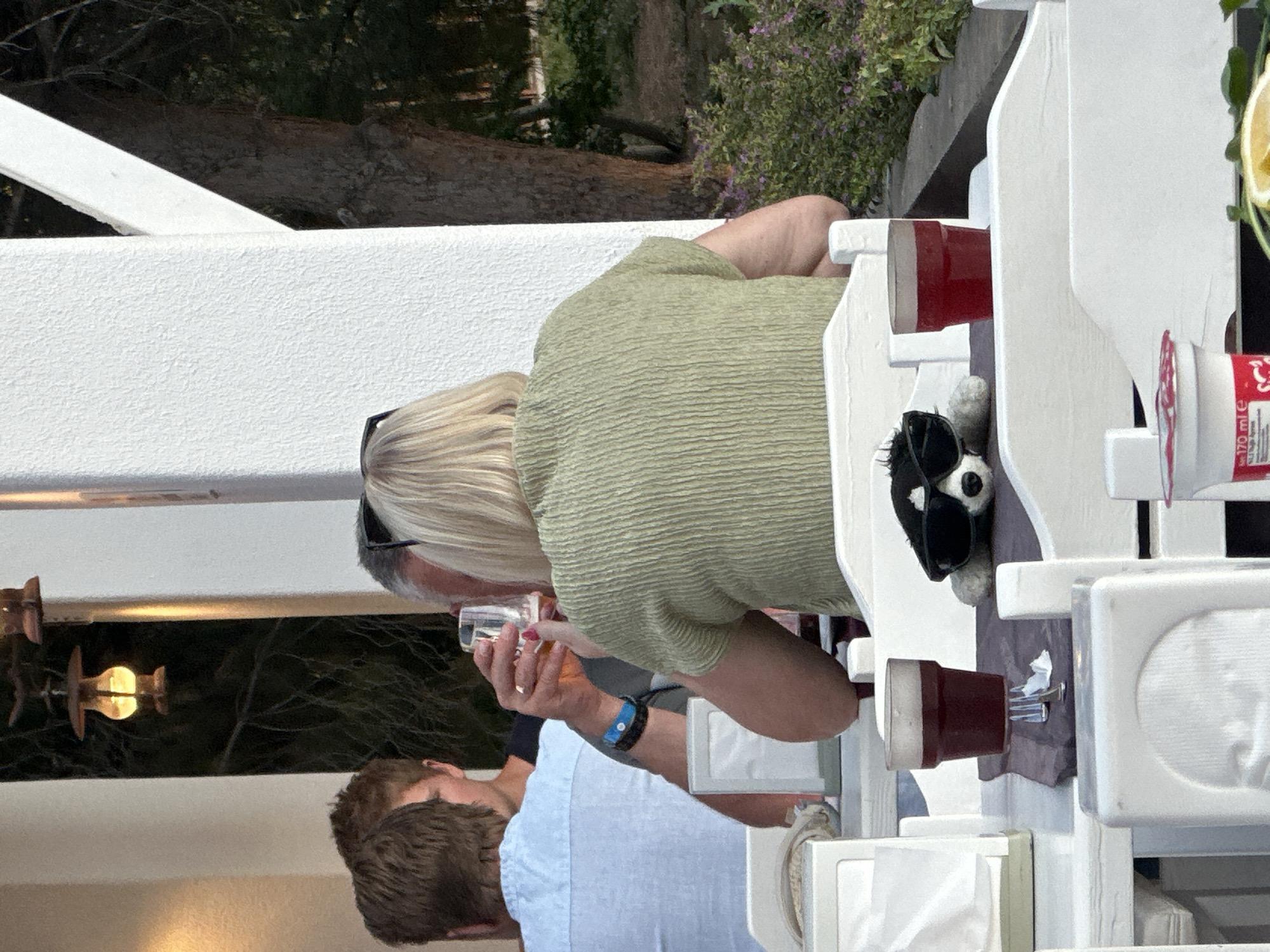 The scene depicts people seated at a white wooden table, likely on a terrace or outdoor dining area in Fethiye, Turkey. A woman with short blonde hair, wearing a light green textured shirt, is drinking from a glass. Another person, wearing a light blue shirt, is visible beside her. A stuffed animal wearing sunglasses is positioned on the table. Red cups and a bottle are also present on the table. The background features trees and vegetation.