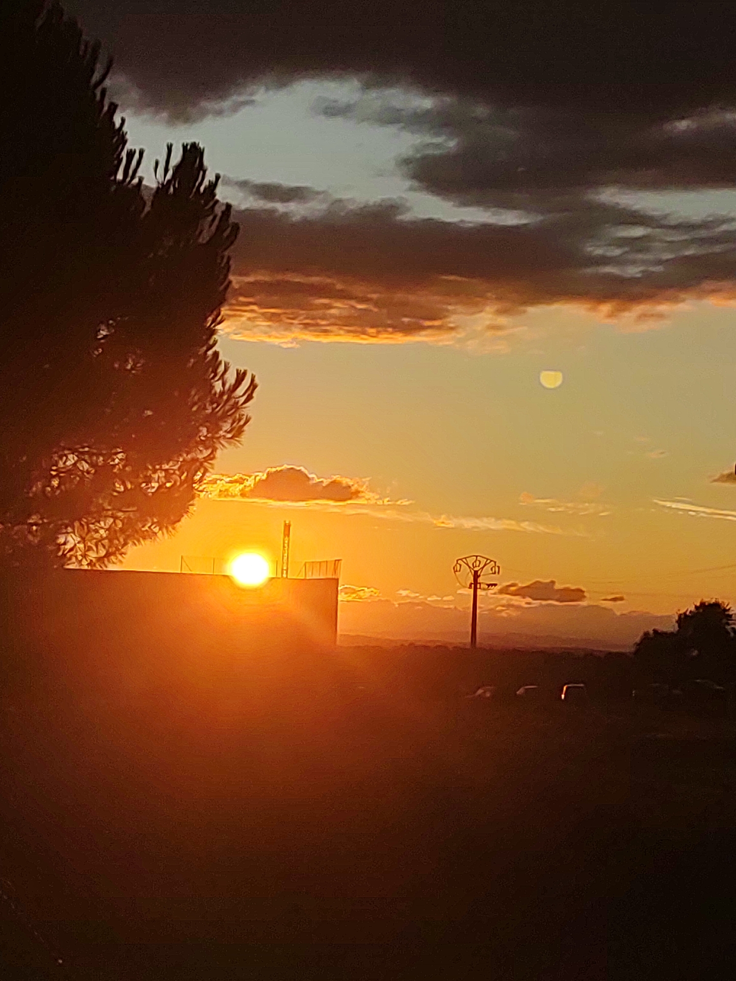 The image depicts a sunset scene with a vibrant orange sky. In the foreground, the silhouette of a large tree is visible on the left. A building with a railing on its roof is situated in the center, with the sun, a bright glowing orb, partially obscured by the building and tree. To the right of the building, a tall, thin utility pole with multiple horizontal crossbars stands against the sky. In the distance, a faint silhouette of mountains can be seen. The sky is a gradient of colors, transitioning from deep gray and dark clouds at the top to bright orange and yellow hues near the horizon. A small, pale crescent moon is visible in the upper right portion of the sky. The ground in the foreground is dark and indistinct. The location is identified as being in Sobradillo, Spain.