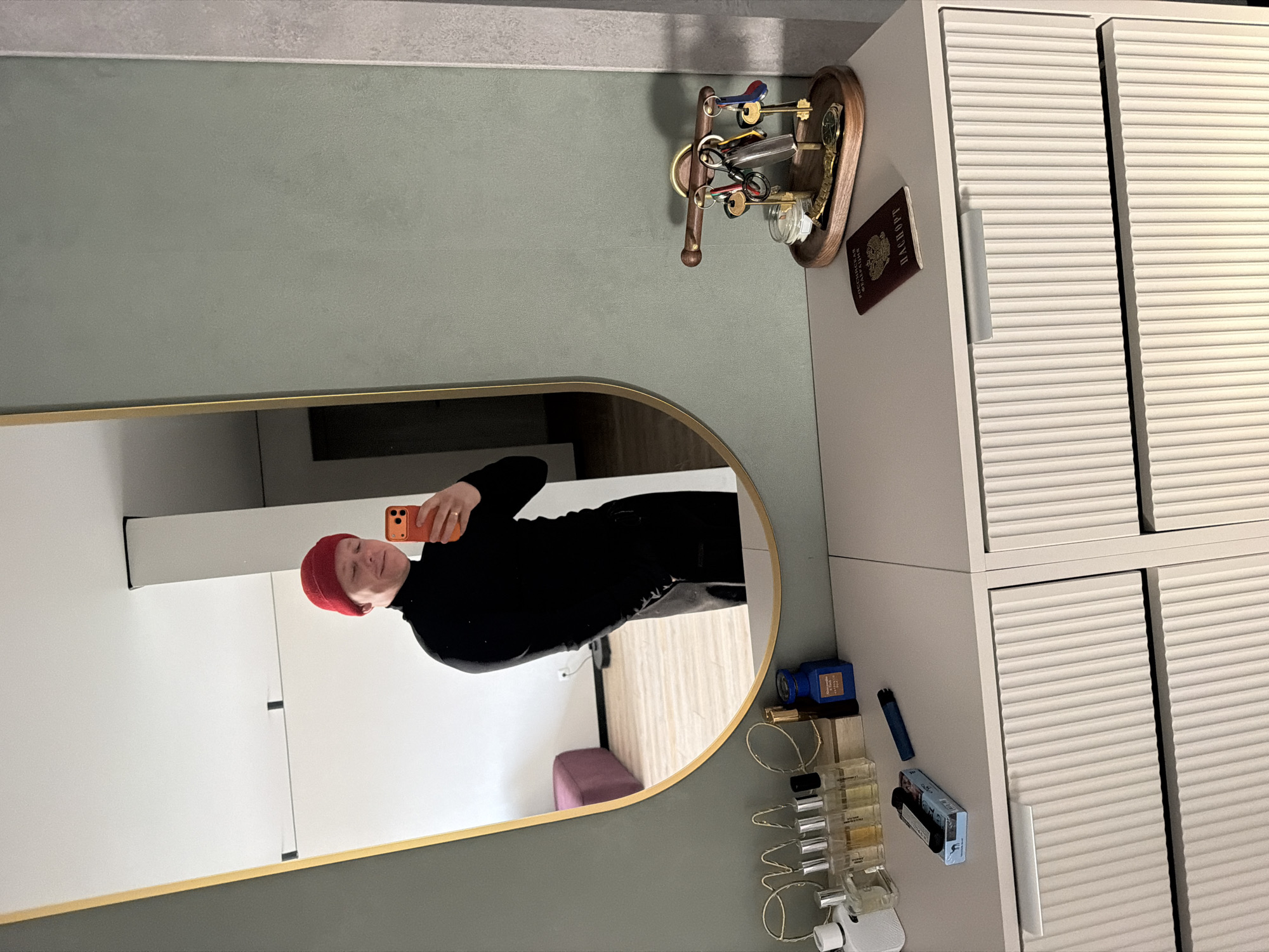A man with a red beanie and black turtleneck is taking a selfie in a mirror. The mirror is located on a wall next to a cabinet. On top of the cabinet are keys, a passport, and some miscellaneous items. Underneath the mirror there are perfume bottles and a blue box. The location is likely an apartment interior in Chaikovsky, Russia.