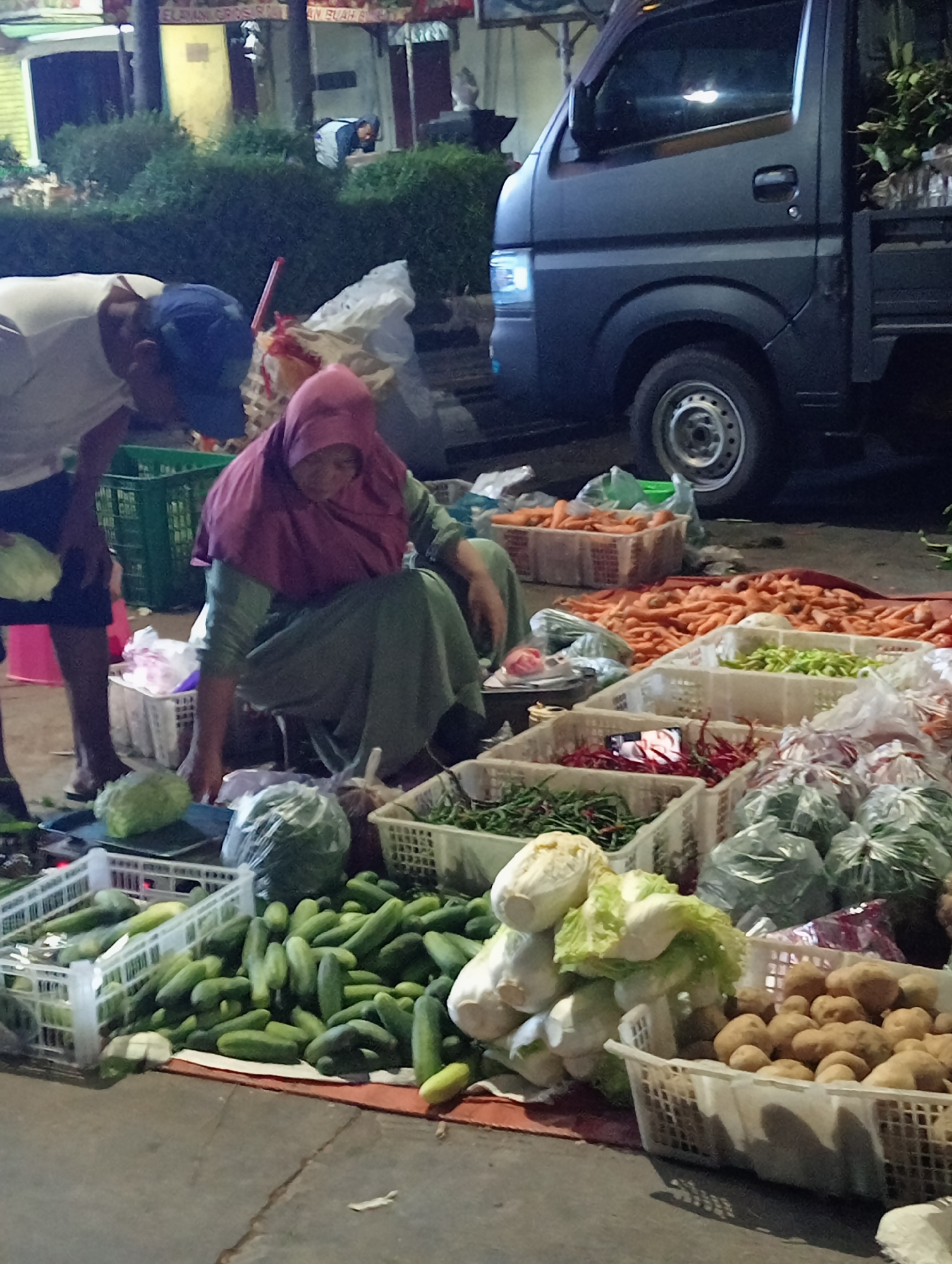 In Demak, Indonesia, an outdoor market scene unfolds with a variety of fresh produce displayed on the ground and in crates. A woman wearing a maroon hijab and a green long-sleeved dress is crouched, appearing to sort through vegetables. To her left, a person in a white t-shirt and blue baseball cap is also bending down, their face obscured. Various vegetables are visible, including a large pile of cucumbers, cabbages, carrots in crates, and bundles of green chilies. In the background, a dark-colored truck is parked, and further back, market stalls with awnings and a building are present, suggesting a busy marketplace environment. The ground is paved, and the lighting indicates it might be evening or early morning.
