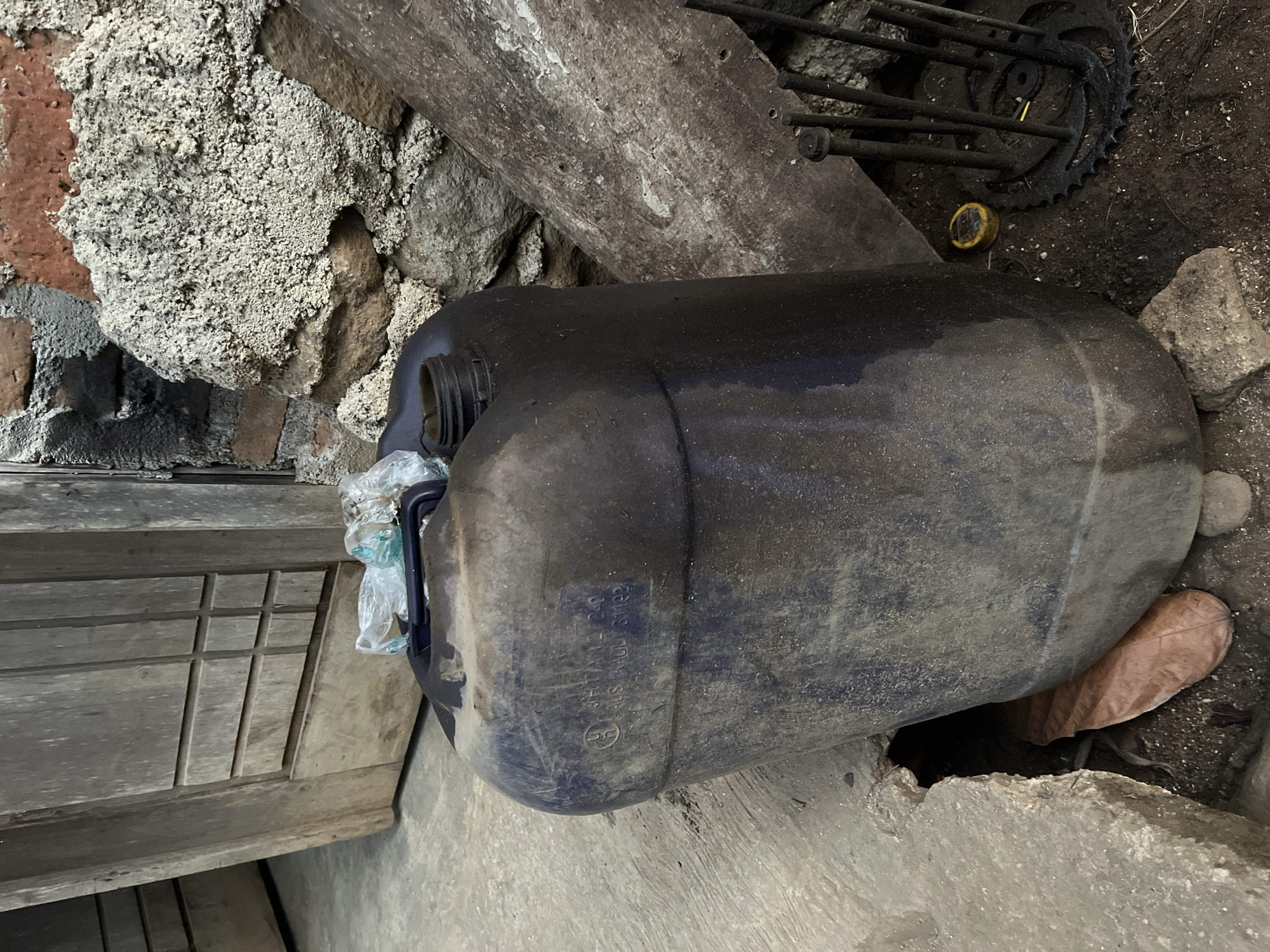 A dusty, dark blue plastic jerrycan sits on a concrete surface outdoors. The jerrycan is mostly filled with a dark liquid and has a black handle. A piece of clear plastic is wrapped around the top of the jerrycan. To the left of the jerrycan is a wooden door with several horizontal and vertical grooves. Behind the jerrycan is a rough stone wall with exposed brick. To the right of the jerrycan are several rusty metal objects, including a cogwheel and some bolts or rods. The ground is a mixture of dirt, gravel, and concrete. The scene appears to be in an outdoor storage or work area, possibly in a backyard or alleyway. The location is specified as Kebumen, Indonesia.