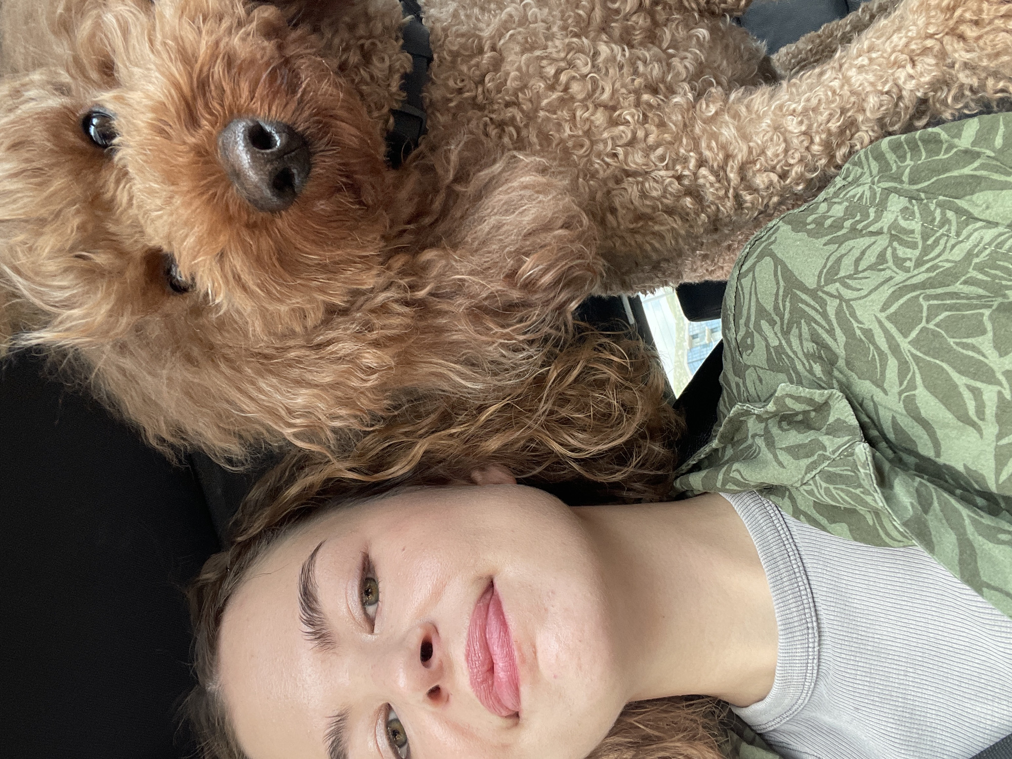 A light-skinned young woman with hazel eyes, light brown curly hair, and pink lipstick is lying on her back in a black upholstered seat. Her tongue is slightly protruding from her mouth. She is wearing a light gray ribbed tank top under a green patterned button-down shirt. Resting on her is a brown Goldendoodle dog. The scene appears to be inside a car. The location cannot be determined from the image.