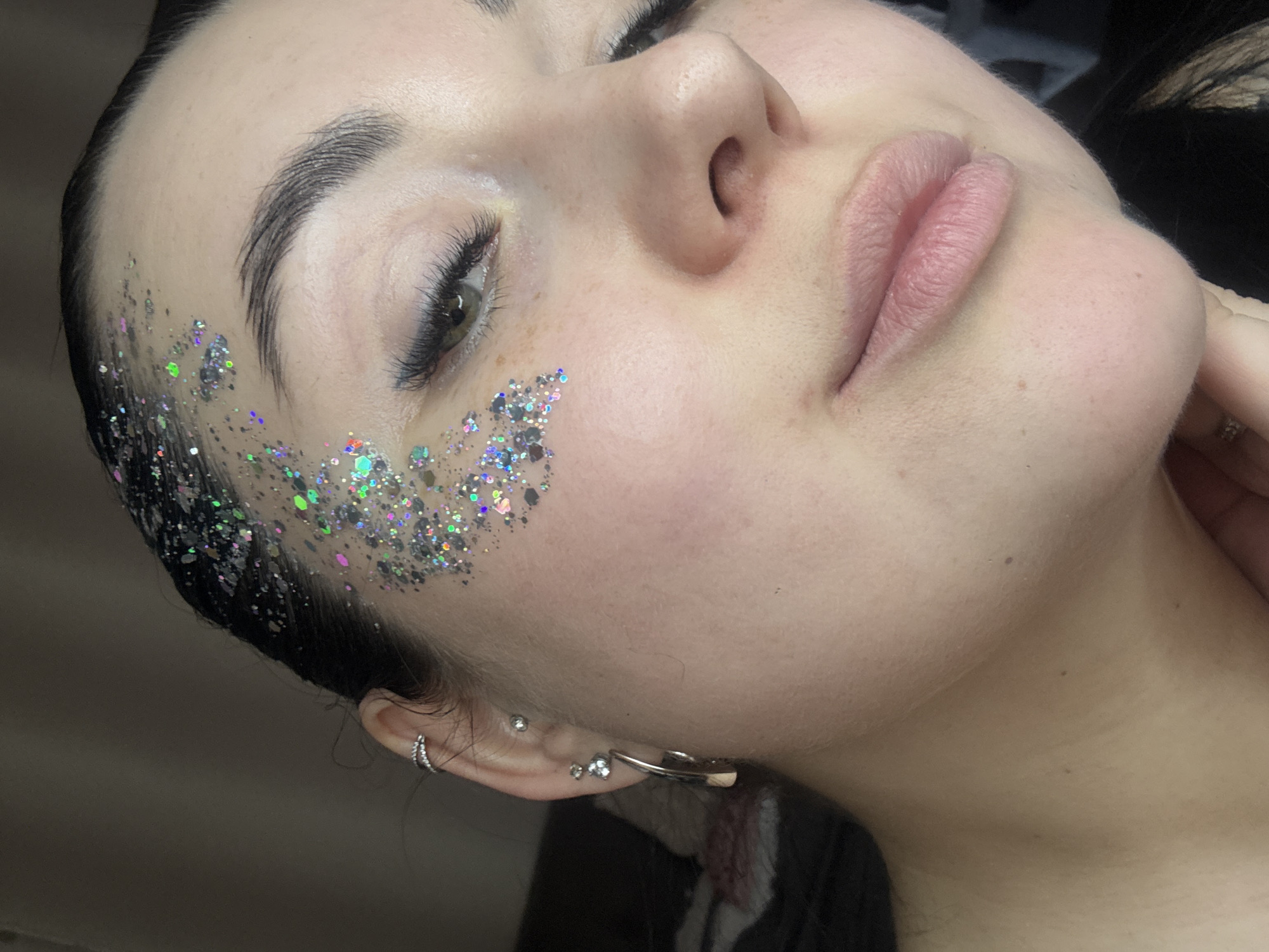 The image is a close-up of a light-skinned person's face, angled upwards to the left. The person has dark hair, dark eyebrows, and fair lips. Their right eye is partially closed, revealing dark eyelashes. Silver and iridescent glitter is applied to the left side of their face, extending from the hairline towards the cheek. Multiple earrings are visible in the person's left ear. The background is out of focus and dark. The location is not discernible from the image alone. There is no indication this image was taken in the city of Chaikovsky, Russia.