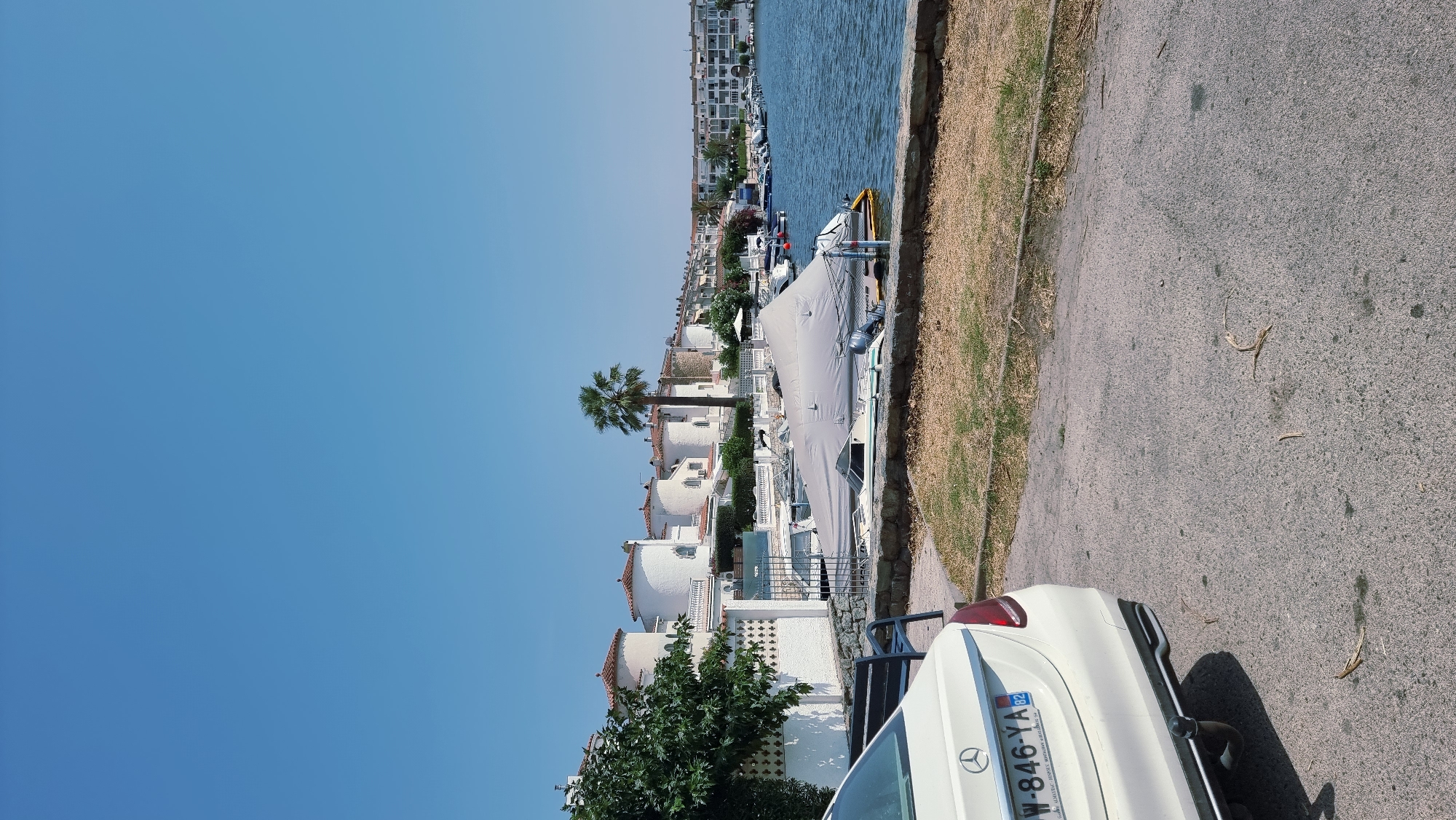 The image captures a view of the Empuriabrava marina in Castelló d'Empúries, Spain, under a clear blue sky. In the foreground, the rear of a white Mercedes-Benz sedan is visible, parked on an asphalt surface. The car's license plate reads "W-846-YA 82". Adjacent to the car, a dark-colored park bench is positioned on a patch of dry grass and dirt.
Beyond the bench, a stone wall borders a strip of dry, light-brown grass. A line of white buildings with distinctive round, turret-like structures and terracotta roofs stretches along the waterfront. A single palm tree stands prominently among these buildings. Further down the waterfront, a row of apartment buildings with balconies faces the water.
The marina itself is filled with numerous boats. Some are covered with grey tarpaulins, while others are uncovered. The water in the marina is calm and reflects the clear sky. The overall scene depicts a sunny day in a coastal town known for its extensive network of canals and marinas.