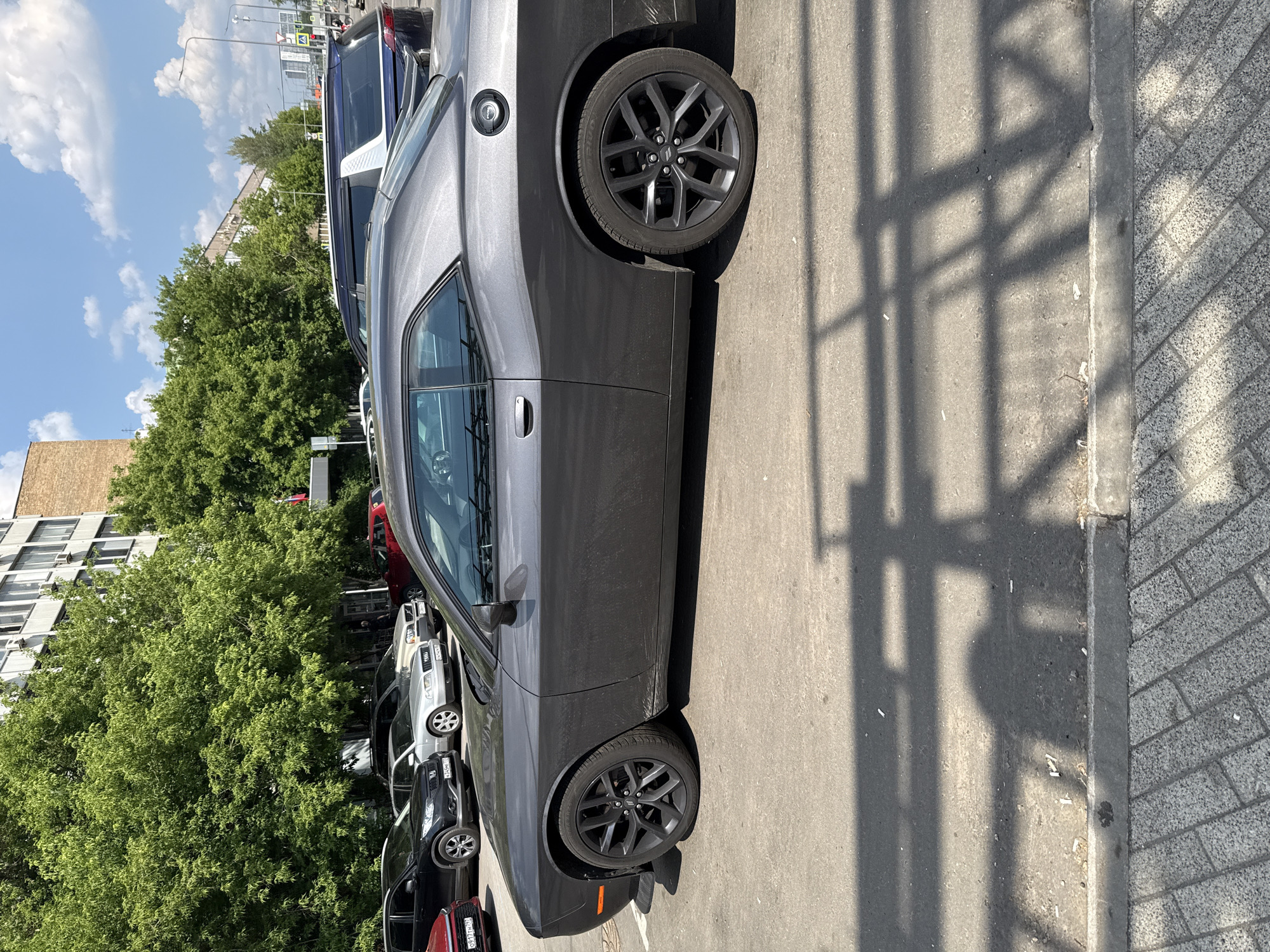 In the city of Khimki, Russia, a grey Dodge Challenger is parked on a paved street in front of a multi-story building. The car is positioned on the right side of the frame, facing left. In the background, several other cars are parked in a line. A large tree with lush green foliage is situated behind the parked cars. The building in the background appears to be an apartment complex or office building. The sky is blue with scattered white clouds. The ground in the foreground is asphalt, with a grey concrete curb separating it from a patterned paving stone area. Shadows from an overhead structure are cast across the asphalt.