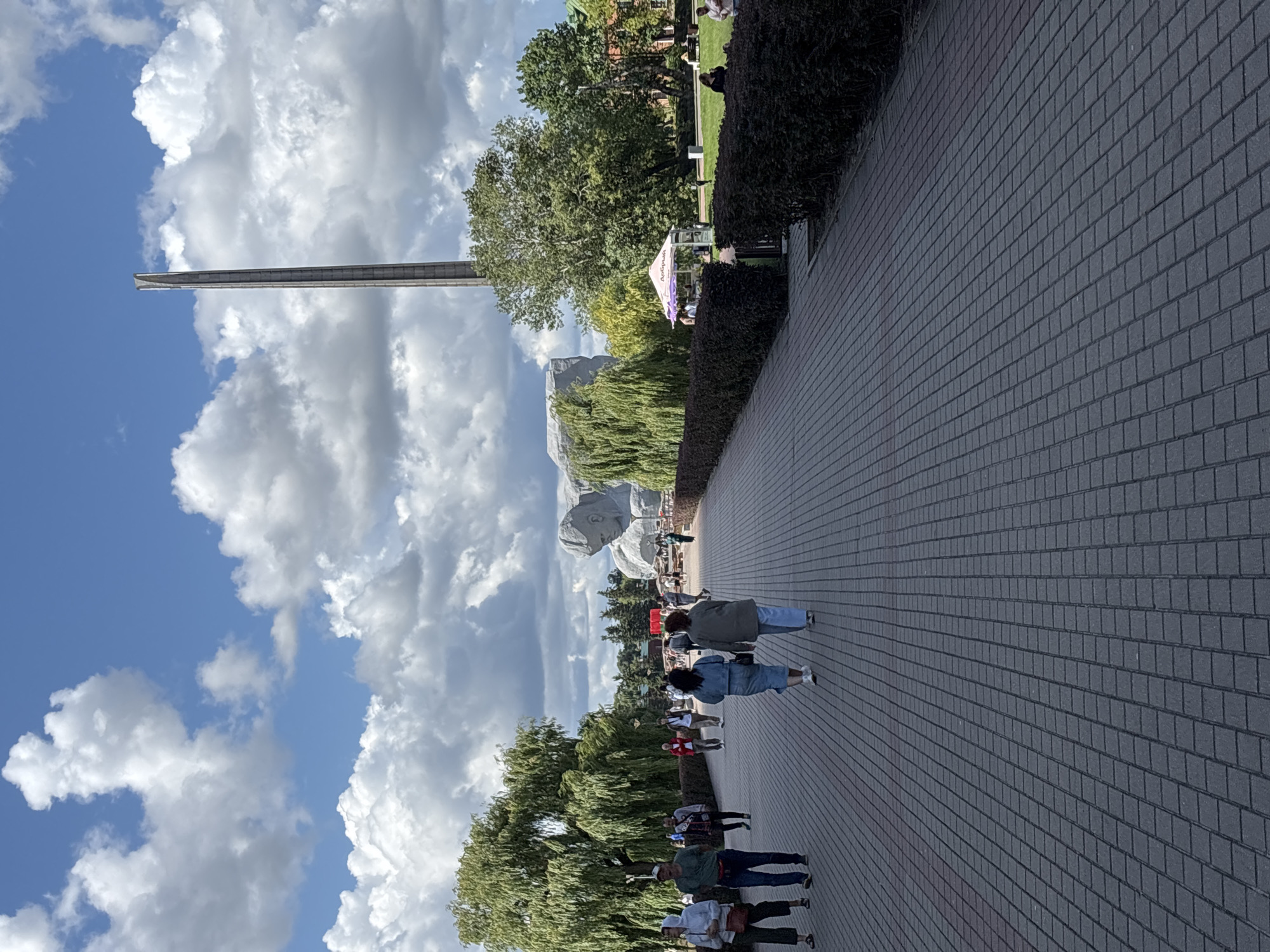 The image depicts a sunny day at the Brest Fortress in Brest, Belarus. A paved walkway, laid with dark gray rectangular stones, leads towards the background, flanked by manicured dark green hedges on the right and trees on the left. Several people are walking on the path, some in small groups, others individually. In the distance, a monumental sculpture of a human head carved from white stone dominates the landscape, with a tall, slender obelisk rising behind it against a bright blue sky filled with fluffy white clouds. A small white tent with purple accents, likely a vendor booth, is set up to the right of the path, near a cluster of trees. Further in the background, green lawns and more trees are visible, along with a red-brick building on the far right. The overall scene suggests a public space with historical significance, attracting visitors on a pleasant day.
