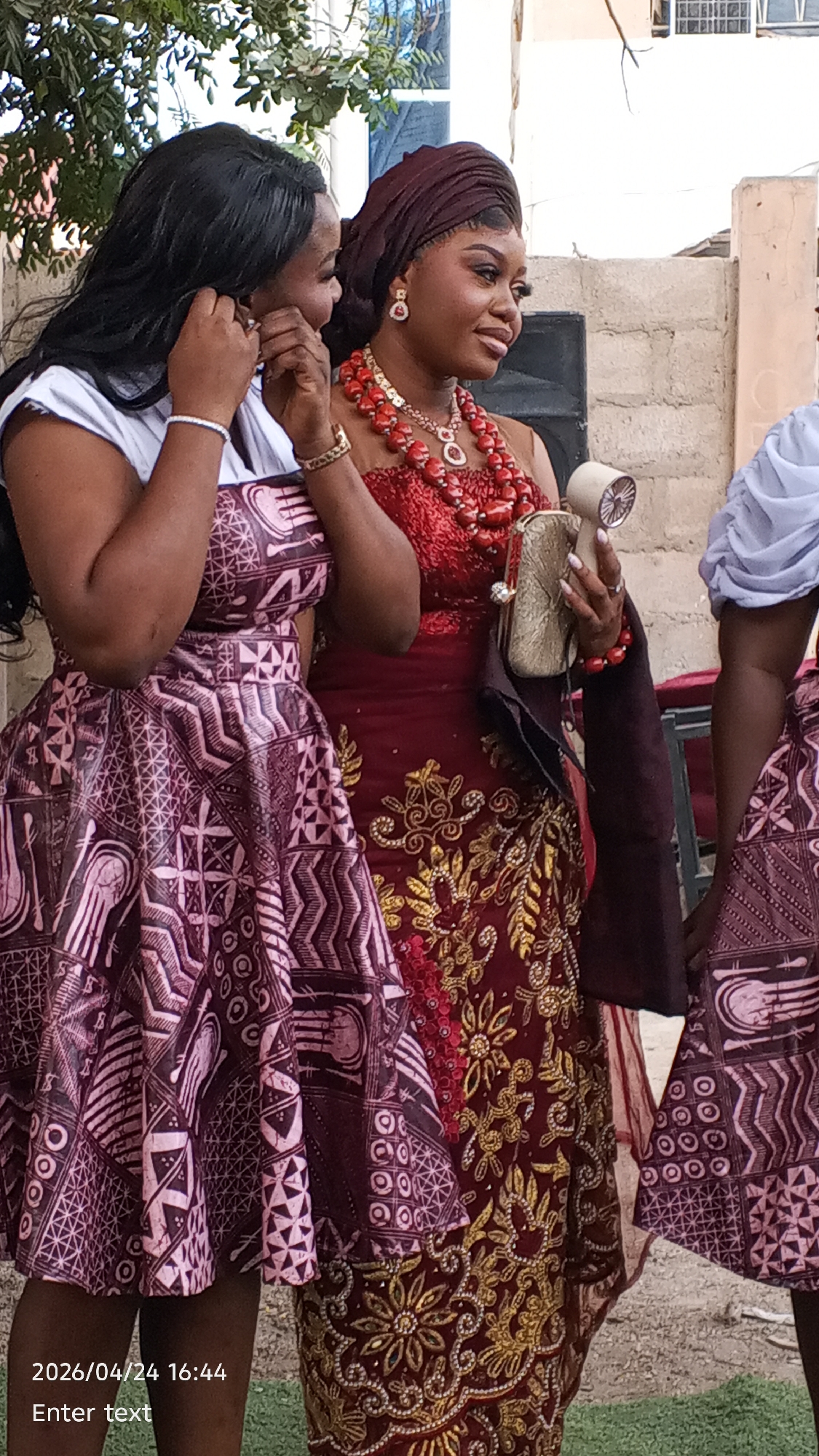 The image captures three women standing outdoors, possibly at an event. The woman on the left is wearing a dress with an intricate geometric pattern, paired with a white top. The woman in the center is wearing a maroon dress adorned with golden floral designs and is accessorized with a red beaded necklace and a headwrap. She is holding a small clutch in one hand and what appears to be a handheld fan in the other. The woman on the right is partially visible, wearing a similar geometric-patterned dress with a light blue top. The environment appears to be a residential or commercial area in the city of Unknown, Unknown, with brick walls and some greenery visible in the background.