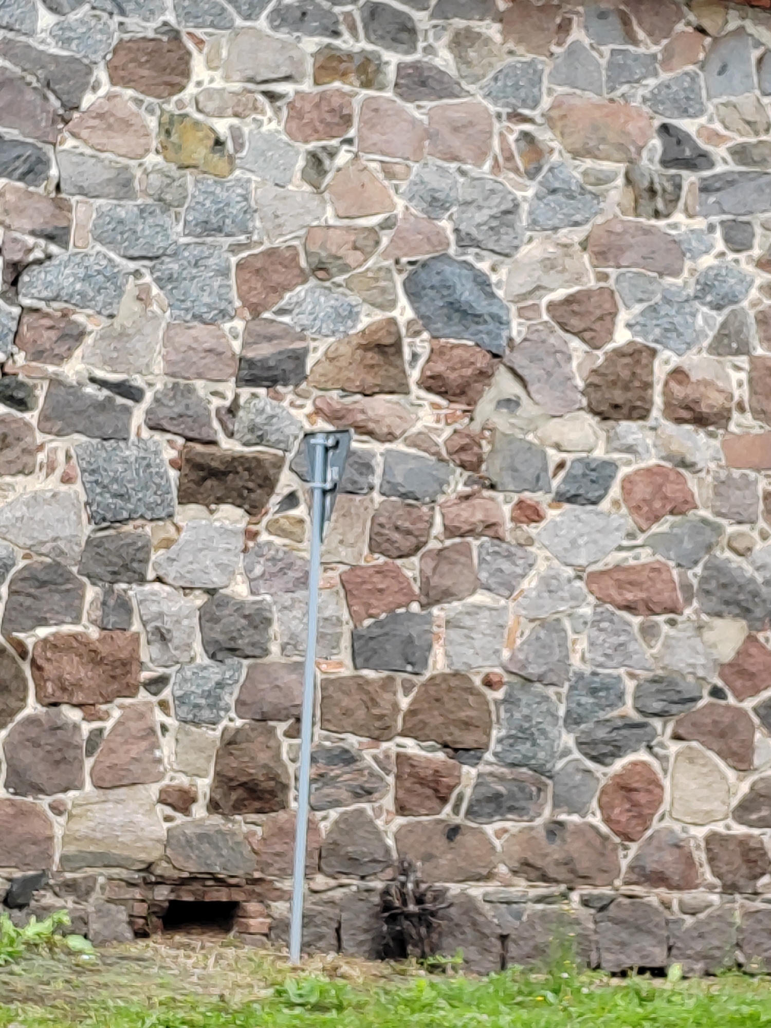 A rustic stone wall, constructed from irregularly shaped stones in various shades of grey, brown, and pink, forms the backdrop of this image. The stones are cemented together with a light-colored mortar, creating a textured surface. A thin, grey metal pole with a small rectangular sign attached is positioned vertically in the center-right of the frame. The base of the wall transitions to a foundation made of smaller, darker bricks, revealing a small, dark opening or recess near the ground. In the foreground, lush green grass and some sparse vegetation create a natural border. The scene appears to be an outdoor environment in the village of Volchynsky Sel'ski Sovet, Belarus, likely part of a historical building or fortification.