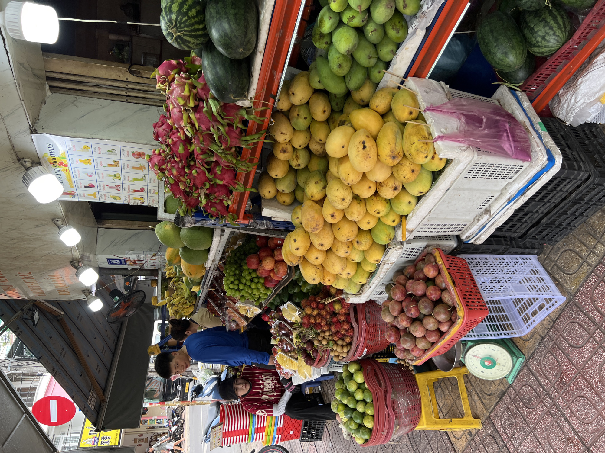 This is a street-level, eye-level, full shot of a fruit stand in Nha Trang, Vietnam. The stand is located on a sidewalk with red and beige patterned tiles. In the foreground, several large red plastic baskets are filled with various fruits, including green limes, rambutan, and passion fruit. To the left, a green mechanical scale sits on the sidewalk. Behind the scale, a young woman wearing a red and white striped shirt and a black beanie sits on a stool, looking towards the camera. She is partially obscured by stacks of colorful plastic baskets.
The fruit stand itself is a multi-tiered structure displaying a wide assortment of fruits. The top shelf holds piles of dragon fruit and watermelons. Below that are stacks of mangoes, both ripe yellow and green varieties, alongside other fruits like grapes and apples. Further down, a selection of sliced fruits such as pineapple and possibly papaya is visible. A poster displaying "Juices and Smoothies" with various fruit illustrations hangs on the wall behind the stand.
Two men are visible behind the counter; one is wearing a blue long-sleeved shirt and appears to be interacting with a customer or preparing fruit, while another person in a light-colored shirt stands behind him. In the background, a sidewalk with more fruit stands and parked motorcycles is visible, along with a red and white "no entry" sign. The overall scene depicts a bustling outdoor market environment.