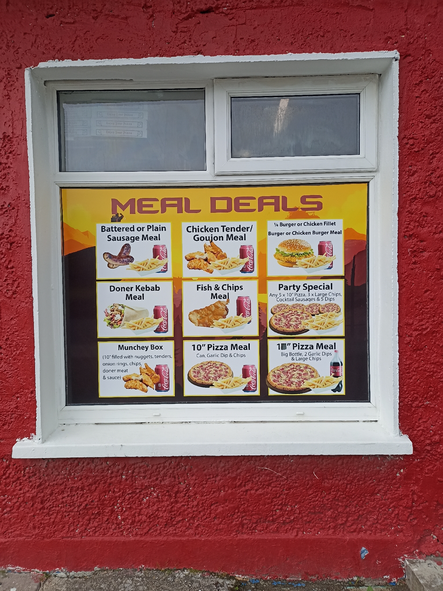 The image shows a bright yellow menu board with various meal deals displayed on a red-painted, textured wall. The menu is behind a window, suggesting it's a takeaway or fast-food establishment. The time of day is not explicitly indicated, but the lighting suggests it could be daytime. There are no people visible in the image.
The menu board features colorful images of food and drinks. The top section prominently displays "MEAL DEALS" in large, stylized letters. Below this, several distinct meal options are presented with accompanying pictures and descriptions. These include:
* **Battered or Plain Sausage Meal:** Depicts sausages and chips.
* **Chicken Tender/Goujon Meal:** Shows chicken tenders and chips.
* **1/4 Burger or Chicken Fillet Burger or Chicken Burger Meal:** Features a burger and chips.
* **Doner Kebab Meal:** Illustrates a doner kebab and chips.
* **Fish & Chips Meal:** Presents a piece of fish with chips.
* **Munchey Box:** Lists the contents including nuggets, tenders, onion rings, chips, doner meat, and sauce.
* **Party Special:** Advertises "Any 5 x 10" Pizza, 3 x Large Chips, Cocktail Sausages & 5 Dips."
* **10" Pizza Meal:** Shows a 10-inch pizza with chips and a drink.
* **Another 10" Pizza Meal:** Displays a 10-inch pizza with chips and a large bottle of Coca-Cola.
Several Coca-Cola branding elements are visible on cans and a bottle, indicating this is likely a fast-food outlet. The overall scene is of a commercial establishment, likely a takeaway food shop, with its menu displayed for customers. The red exterior paint and the style of the window suggest a potentially older building. The location context of Annagry, Ireland, implies a local eatery in a coastal or rural setting.