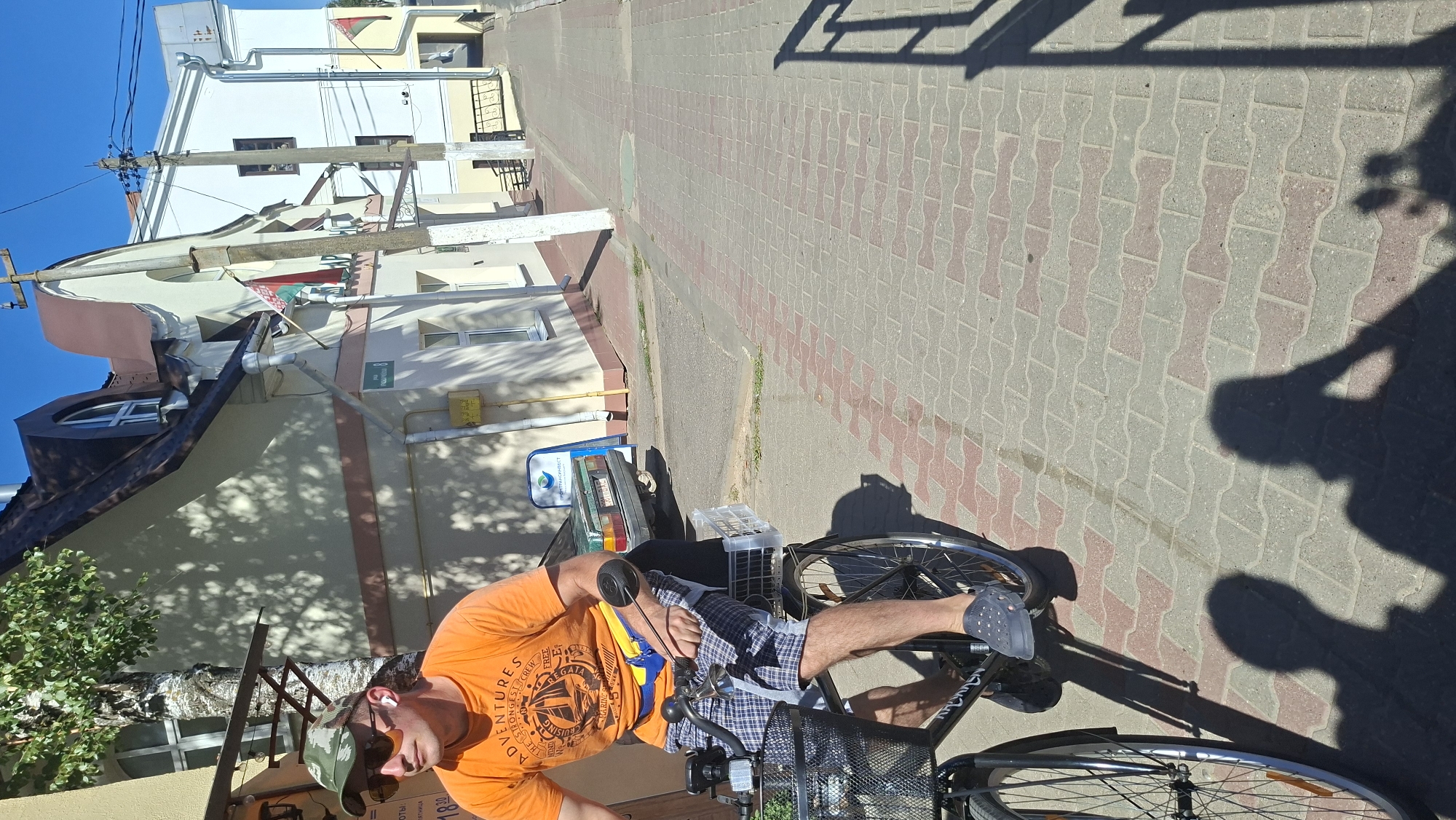 A man wearing a camouflage hat, sunglasses, and an orange t-shirt with a graphic print is riding a bicycle. He has a yellow and blue fanny pack around his waist and is wearing plaid shorts and grey clogs. The bicycle has a black basket in front and a white plastic crate behind the seat. The man is on a brick-paved street in Pastavy, Belarus. Behind him are buildings with cream-colored walls and brown roofs. A green car is parked on the side of the road. The sun is shining, casting shadows on the ground.