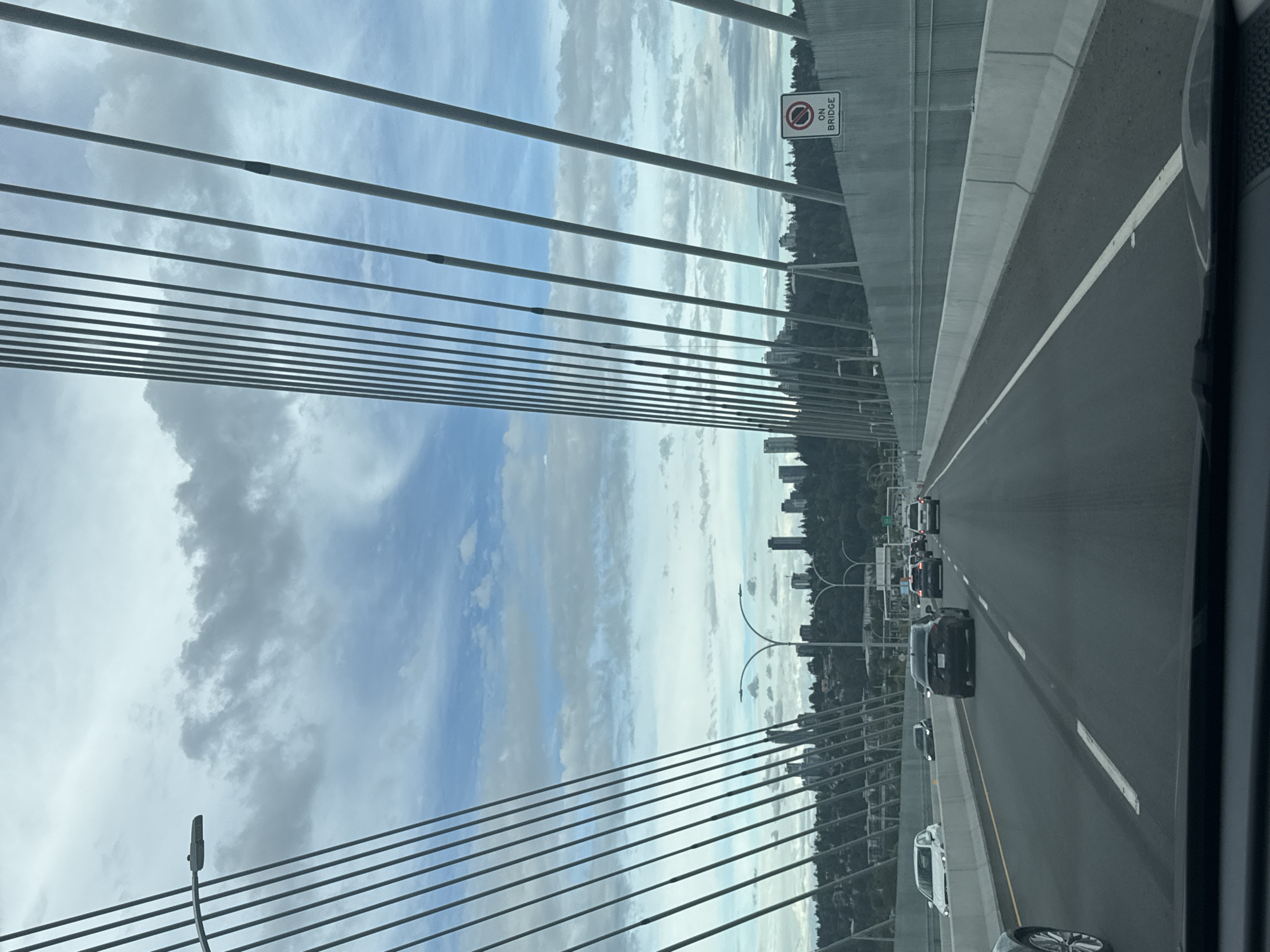 The image shows a view from a vehicle traveling on a multi-lane highway, likely a bridge, with large suspension cables visible on either side. The sky is partly cloudy, with patches of blue and grey. In the distance, a city skyline with several tall buildings is visible against a backdrop of green, tree-covered hills. Traffic is moving in the same direction as the viewer's vehicle, with several cars on the highway. A road sign with a red circle and a diagonal line, indicating a prohibition, and the text "ON BRIDGE" is visible on the right side of the highway. The highway itself is paved with asphalt and marked with white lane lines. The concrete barriers and the overall infrastructure suggest a modern, well-maintained road. The scene is consistent with a major transportation artery in a developed urban or suburban area. Based on the skyline and bridge architecture, this location is likely the Patullo Bridge in Surrey, Canada, connecting Surrey to New Westminster.