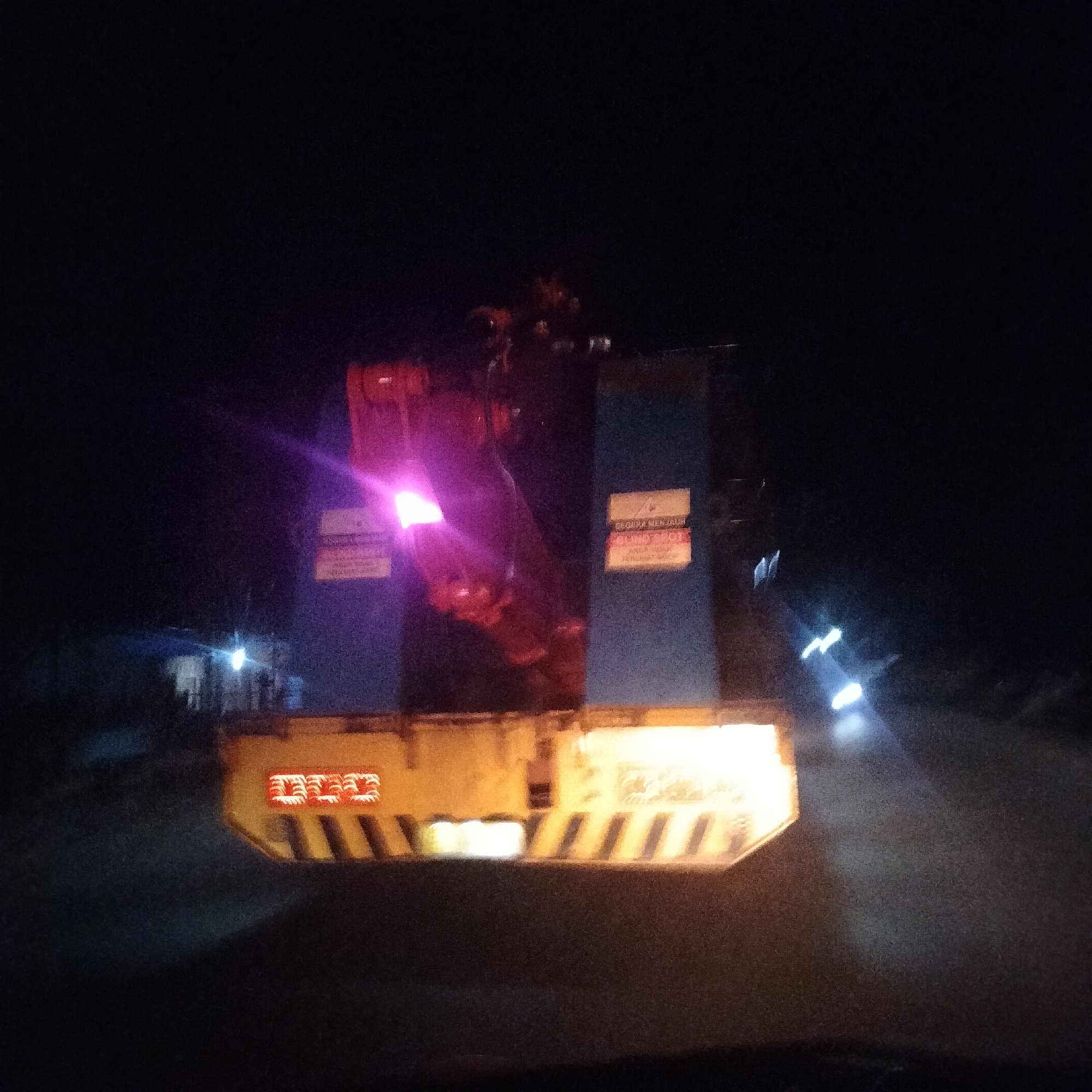 At night in Teupinbatee, Indonesia, the rear of a yellow and blue heavy machinery transport vehicle is visible on a dark road. The vehicle has lights illuminated and warning signs posted on its back. The surrounding environment is dark, with blurred lights in the distance suggesting other vehicles or structures. The road surface is dark and appears to be wet or reflective.