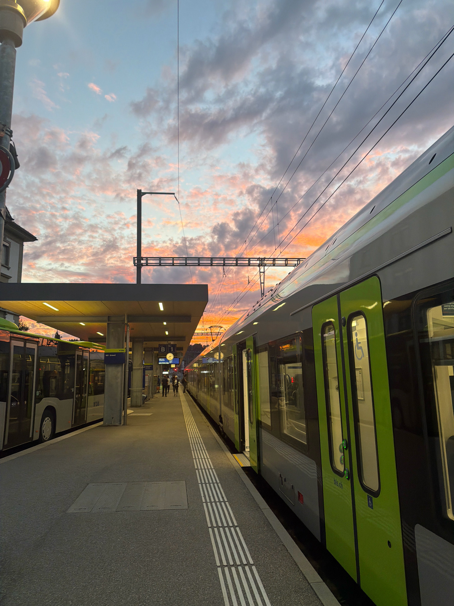 A train station platform in Huttwil, Switzerland, is depicted at dusk. A green and silver train occupies the right side of the frame, with its doors open. To the left, a bus is parked at the platform. The platform itself is paved and marked with tactile paving for the visually impaired. A covered waiting area with lighting extends over the platform. The sky above is filled with dramatic clouds illuminated by the setting sun, casting warm hues of orange, pink, and purple. Overhead power lines for the train are visible against the colorful sky. A few individuals are walking on the platform in the distance. The scene captures a moment of transition at the end of the day, with public transportation at rest and the sky ablaze with color.
