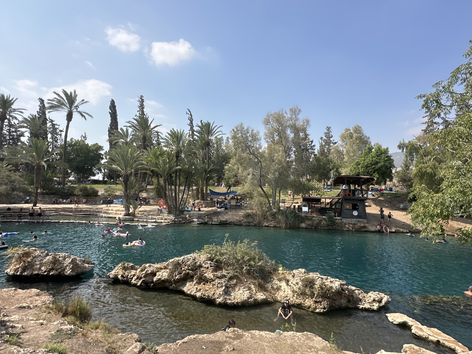 This is a wide shot of a natural hot spring located in the city of Emek HaMaayanot Regional Council, Israel. The water is a clear turquoise color and is surrounded by rocky terrain and lush greenery, including numerous palm trees. Several people are visible in the water, some swimming and others relaxing on inflatable tubes. On the banks of the spring, there are stone steps, seating areas, and a wooden structure that appears to be a lifeguard station or changing room. The sky is clear blue with a few scattered white clouds.