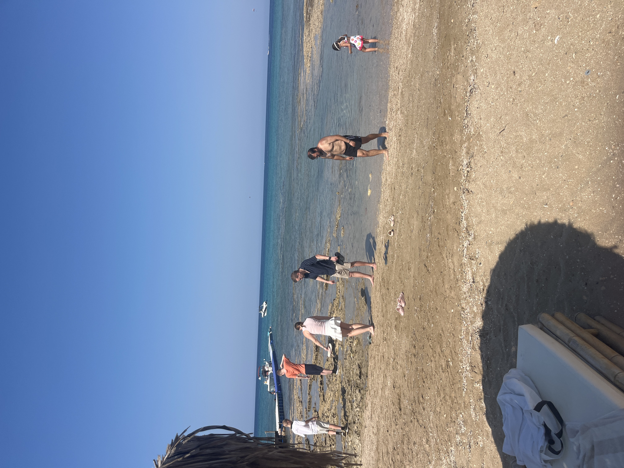 The image depicts a beach scene in Protaras, Cyprus, under a clear blue sky. In the foreground, a thatched umbrella casts a shadow over a white mattress with a bamboo base, with a white shirt draped over it. The sandy beach is strewn with shells. Several people are present. On the left, two young men stand on the shore, one in a white t-shirt and shorts, the other in an orange t-shirt and dark shorts. Further back, a woman in a light pink top and white skirt is looking down, and a man in a striped shirt and khaki shorts stands next to her. In the shallow water to the right, a man in black swim trunks walks, and a young girl in a patterned swimsuit and hat stands, looking at her feet. In the background, a white pier extends into the turquoise sea, with two boats docked and another boat further out. A faint parasail can be seen in the far distance.