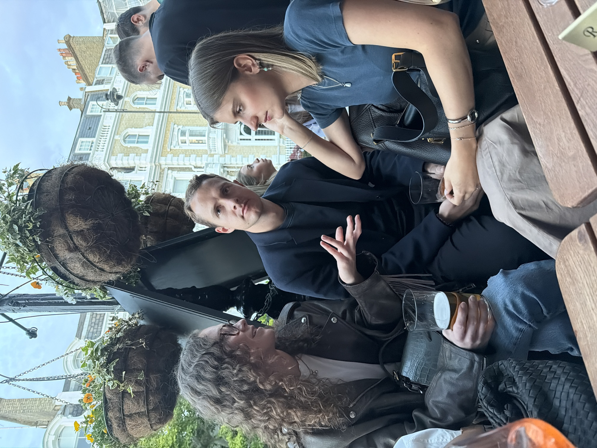 A group of people are sitting at an outdoor table at a pub in London, United Kingdom. The foreground features a woman with curly hair wearing glasses and a leather jacket, holding a glass of beer. A man sits next to her in a dark blue jacket, gesturing with his hand. Above them, a woman in a blue shirt looks down. Several people are crowded around the table. The table is cluttered with bags and drinks. Hanging flower baskets decorate the area. In the background are classic London townhouses with white facades and detailed architectural features. The sky is overcast.