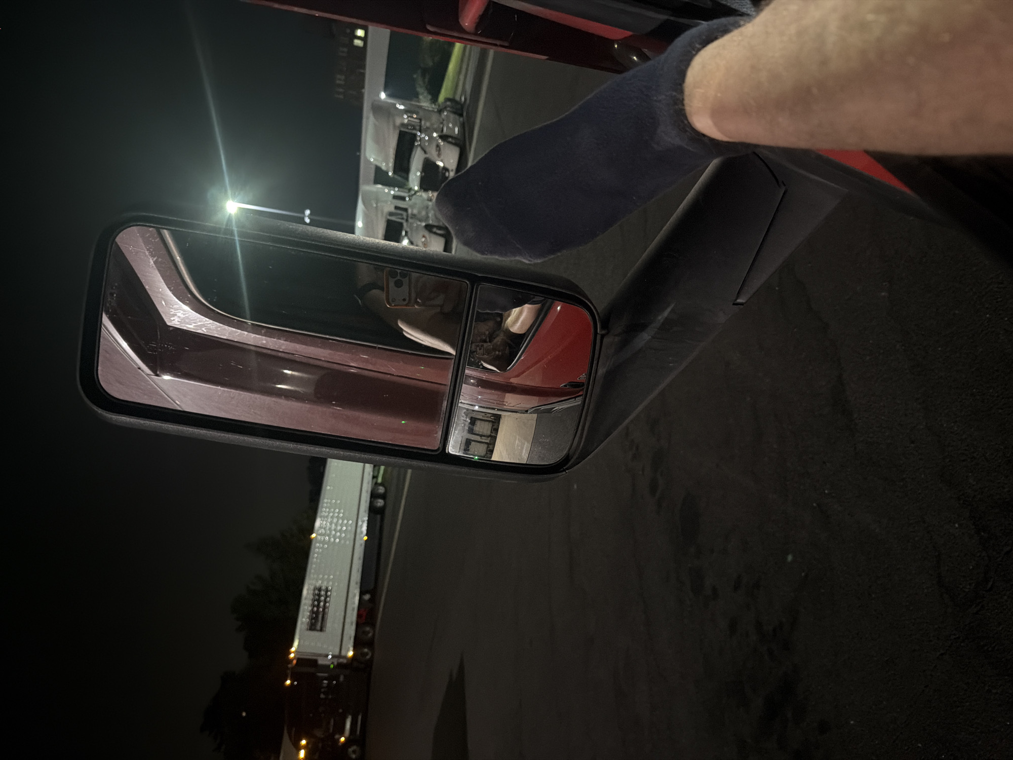 At night, a view from the interior of a semi-truck shows a large side mirror reflecting a person's arm and hand, which is partially covered by a dark blue sock. The arm is extended out of the truck window. In the background, several other semi-trucks are parked. The scene appears to be a truck stop or parking lot. The location is New Britain, Connecticut, United States of America.